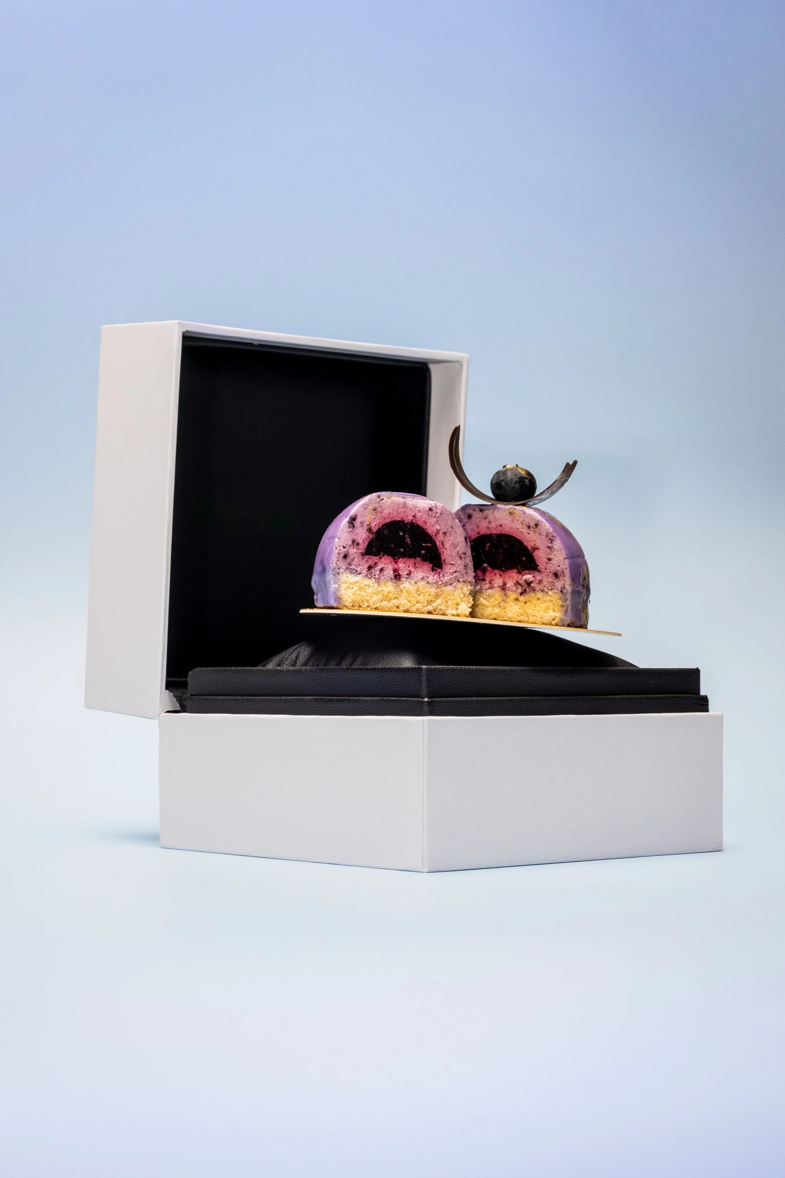 Luxury food photography of sliced berry mousse cake with chocolate garnish in premium white gift box against soft blue backdrop.