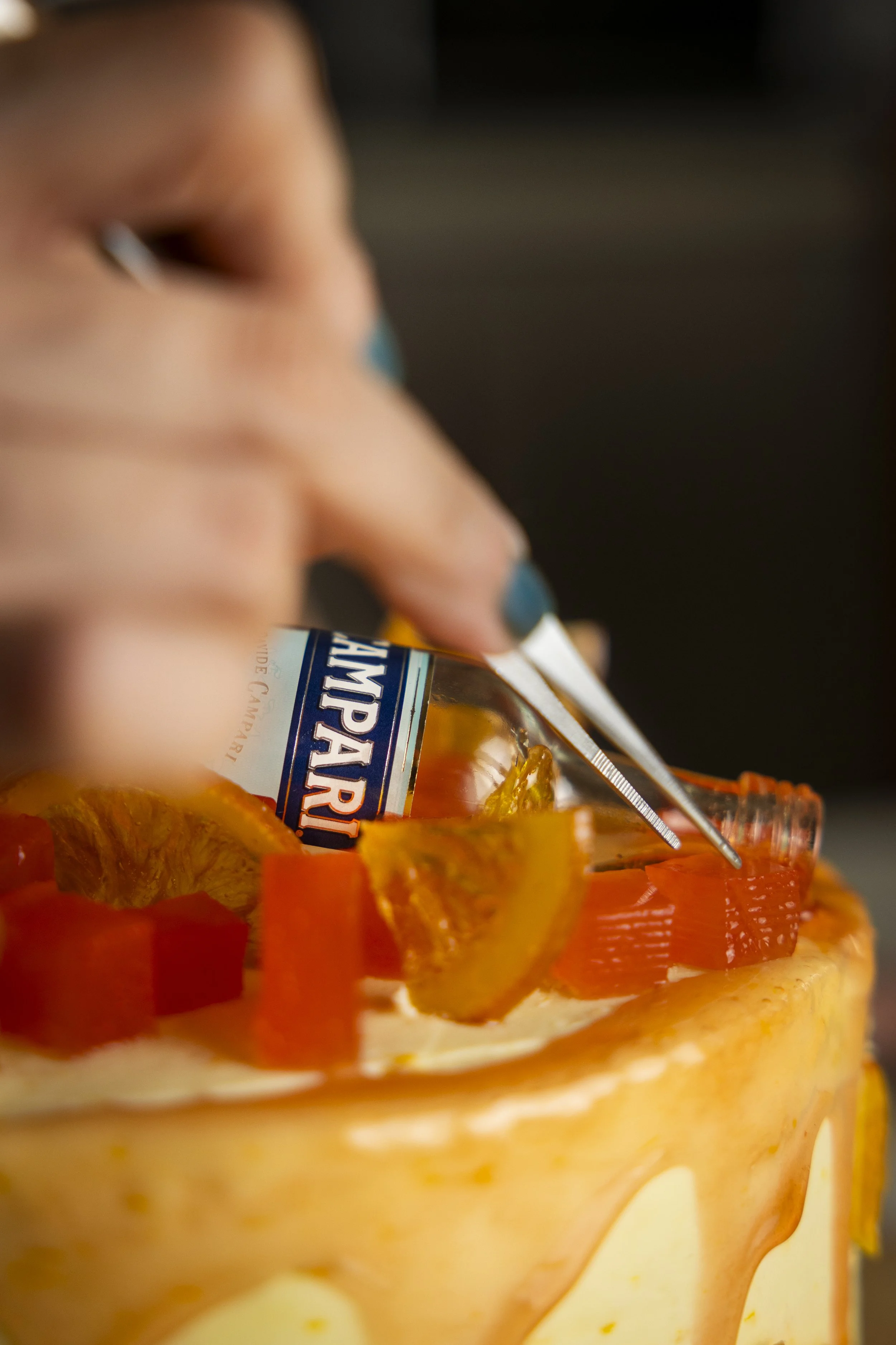 Food photography behind the scenes of Campari branded cake styling with tweezers, candied citrus and caramel drip decoration.