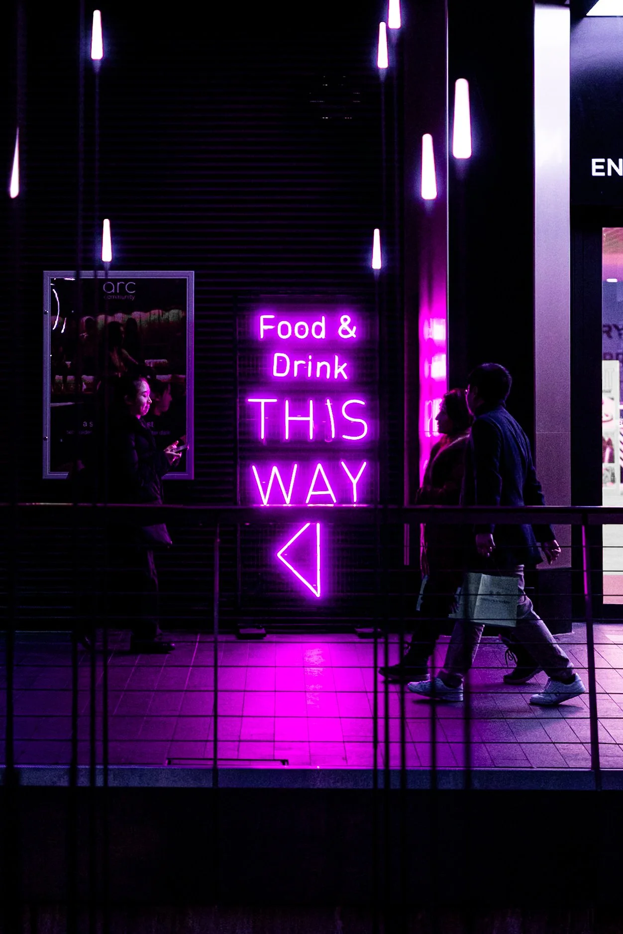 Night urban photography of pedestrians beside glowing magenta neon Food and Drink This Way sign with vivid purple reflections in dark shopping interior.