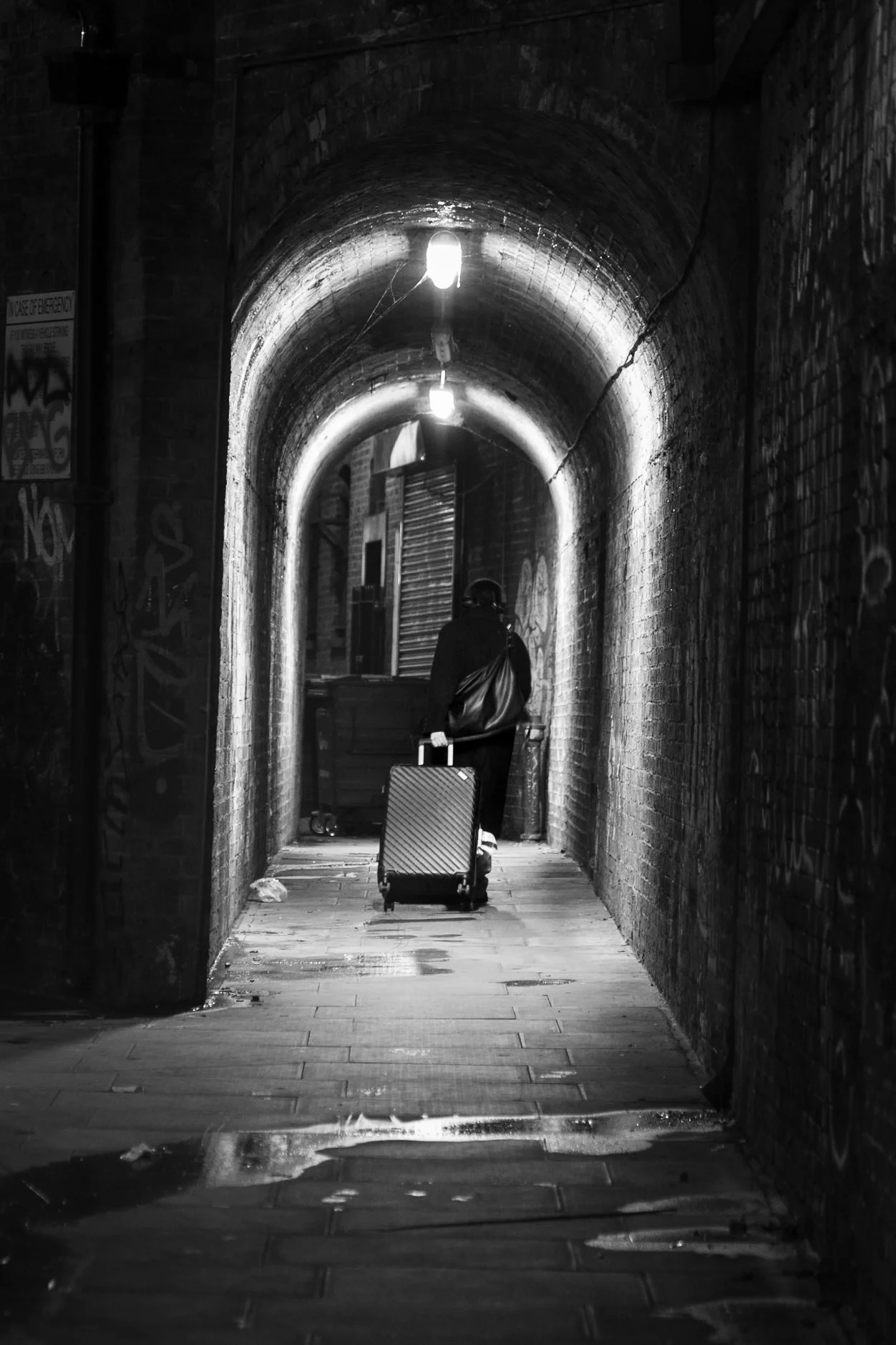 Black and white London night street photography of lone figure with suitcase in Victorian brick arch tunnel with graffiti and wet flagstones.