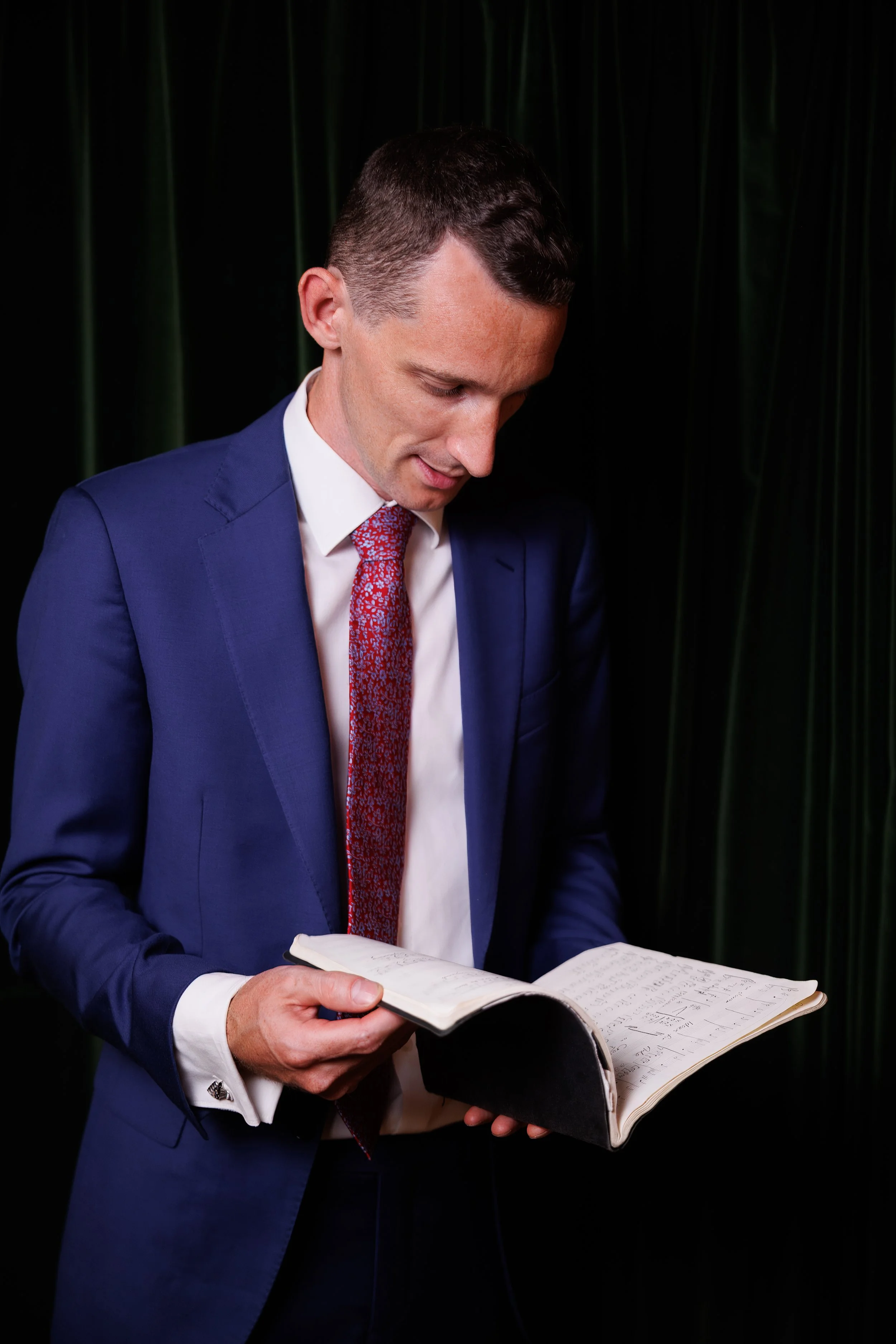 Professional classical musician portrait of conductor in blue suit studying musical score against dark green velvet studio backdrop.
