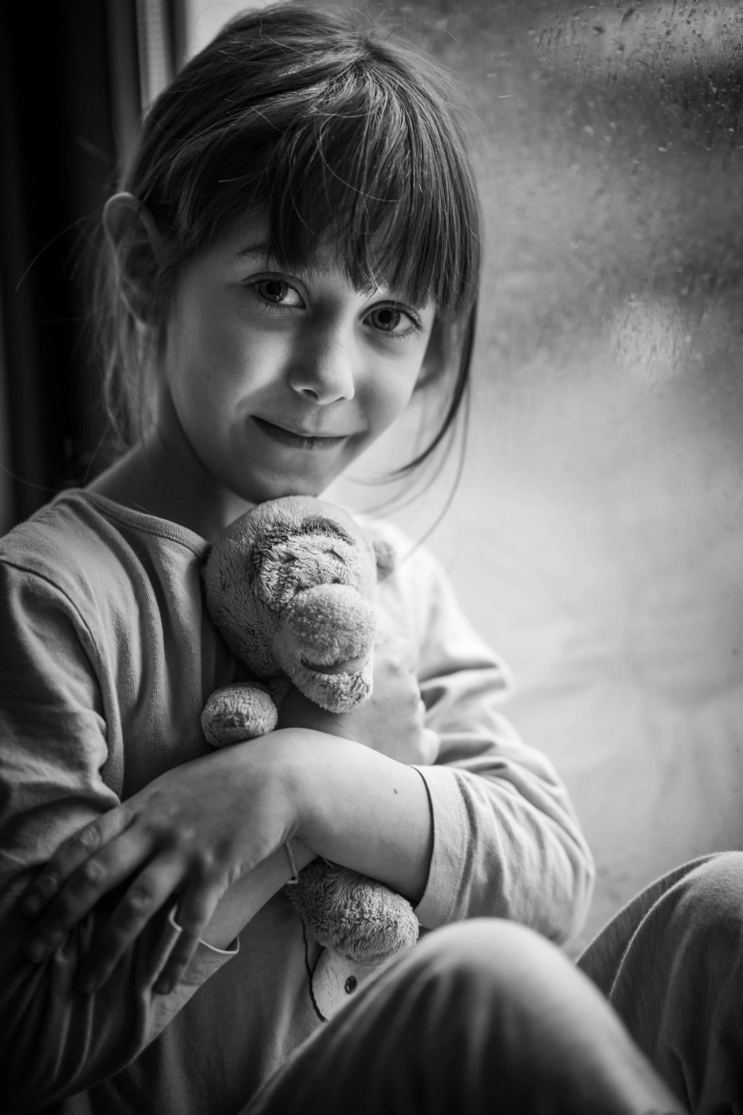 Emotive black and white childhood portrait of young girl hugging teddy bear by rainy window in natural light photography.