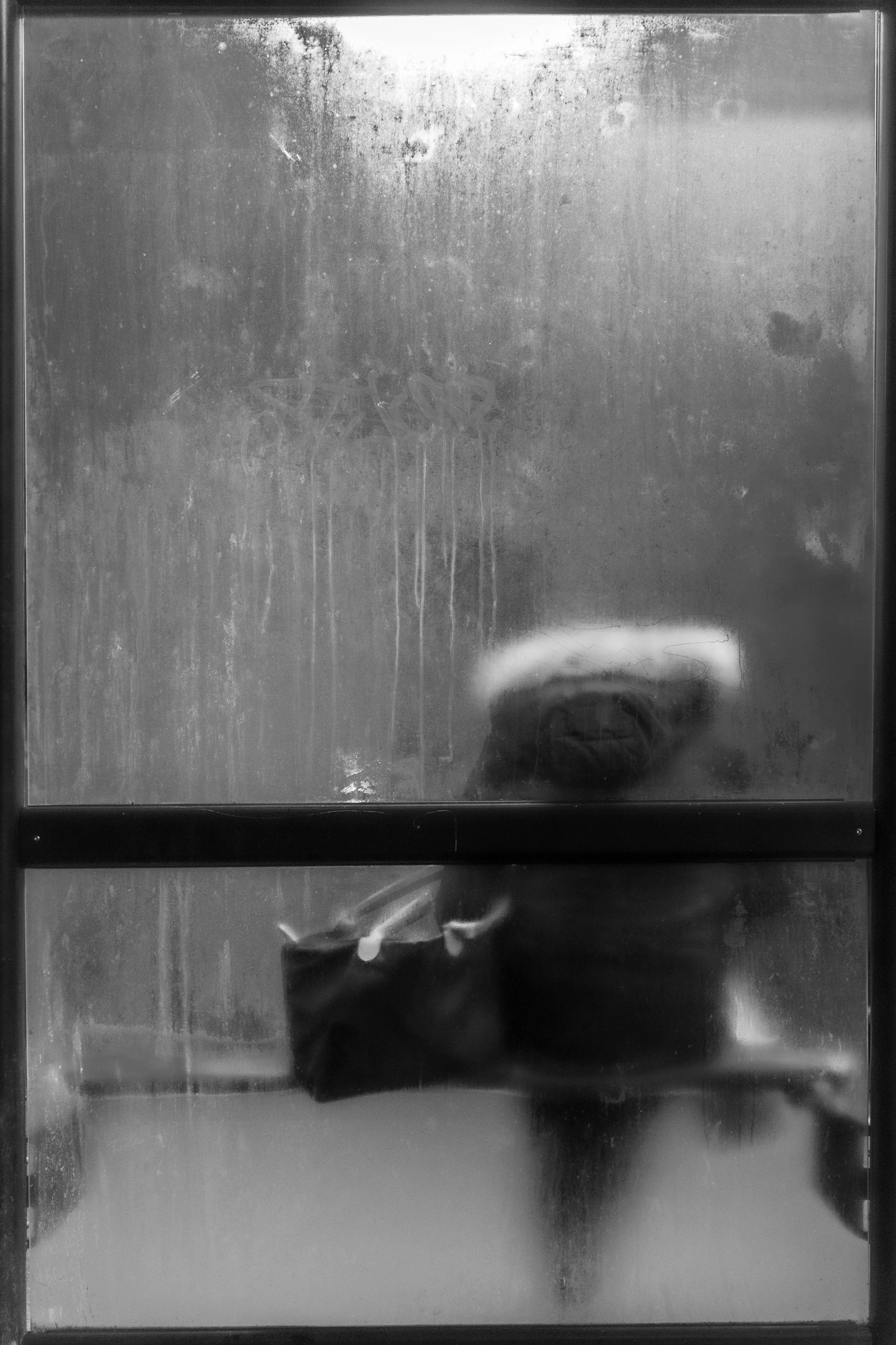 Moody monochrome street photograph of ghostly blurred woman with bag seen through dirty fogged double window with streaked condensation.