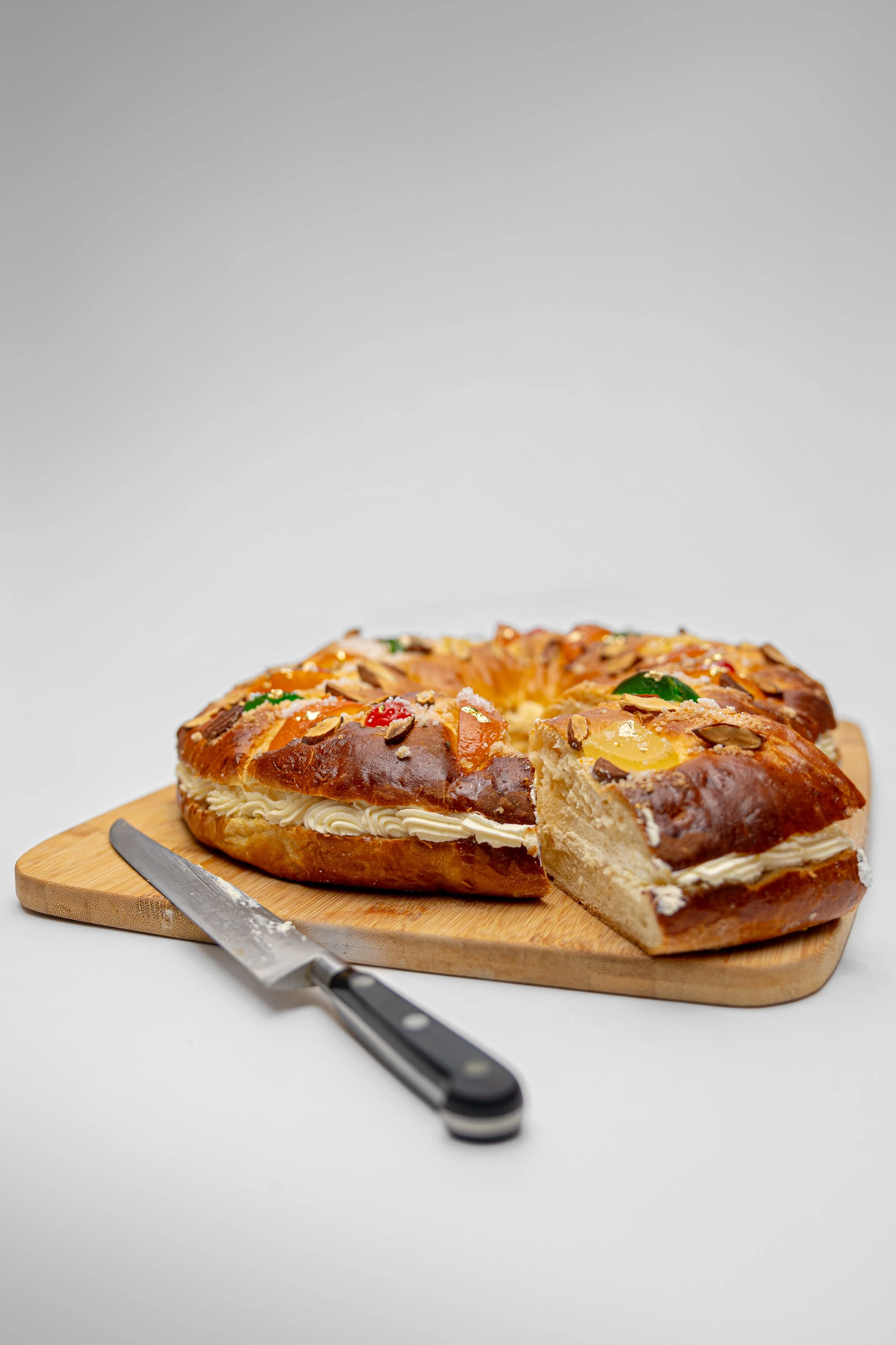 Commercial food photography of traditional Roscón de Reyes Spanish Kings cake with candied fruit, flaked almonds and cream filling on wooden board against white background.