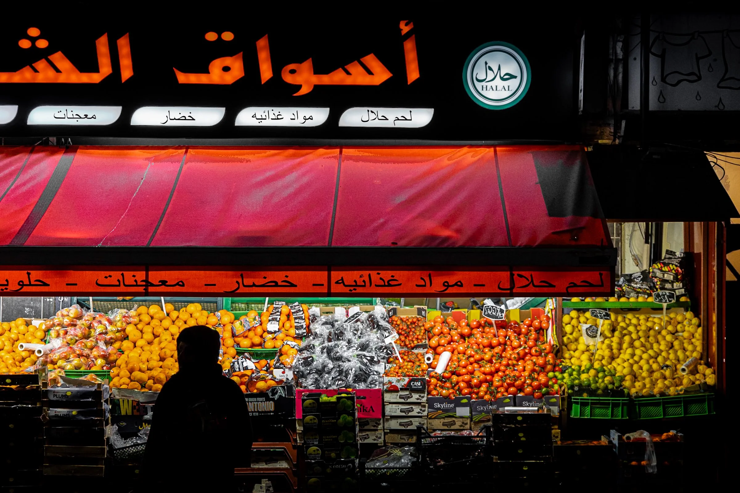 Night street documentary photography of halal market stall with Arabic signage, silhouetted figure and colourful fruit display in urban setting.