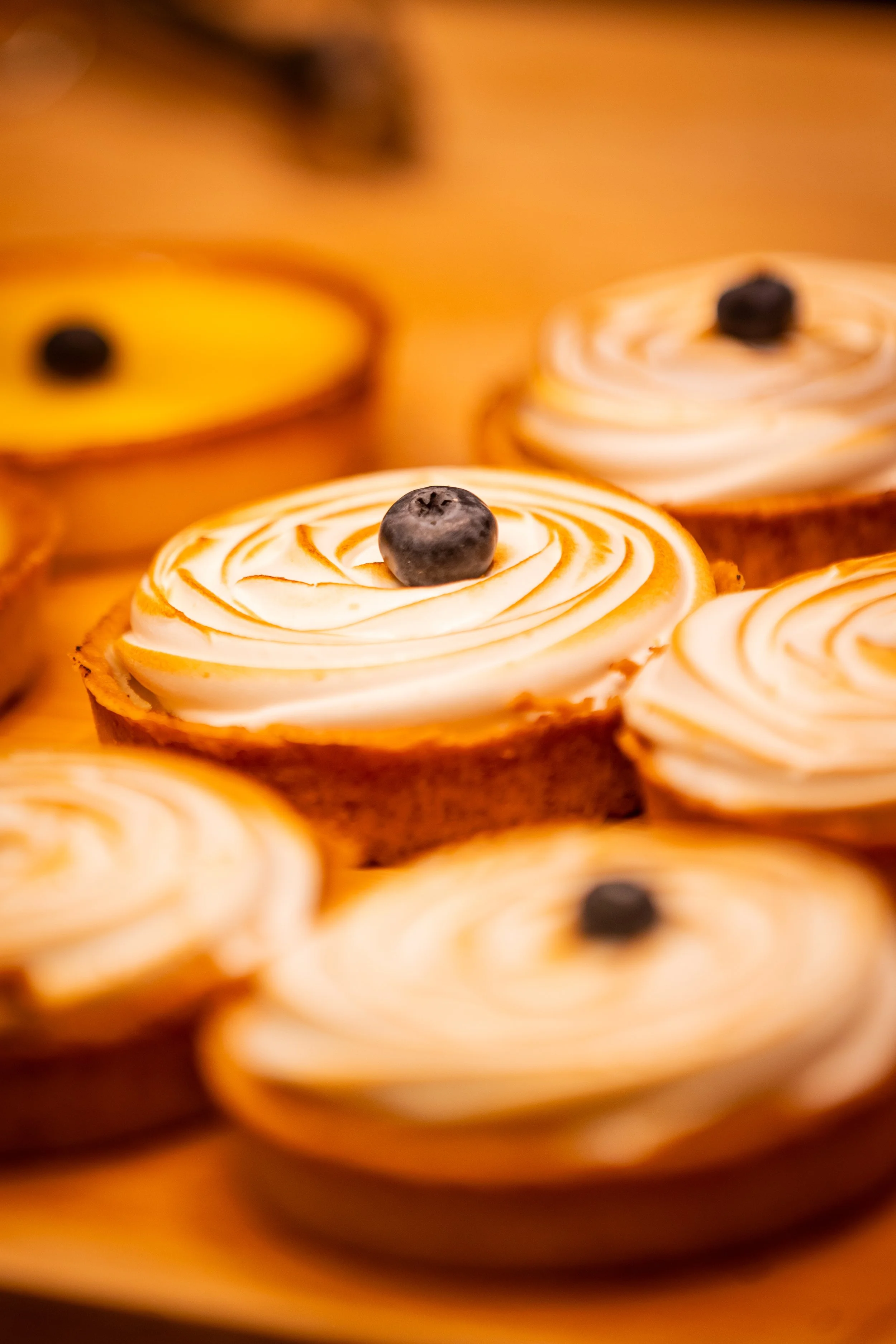 Commercial food photography of lemon meringue tarts with toasted spiral topping and blueberry garnish in warm golden bakery setting.