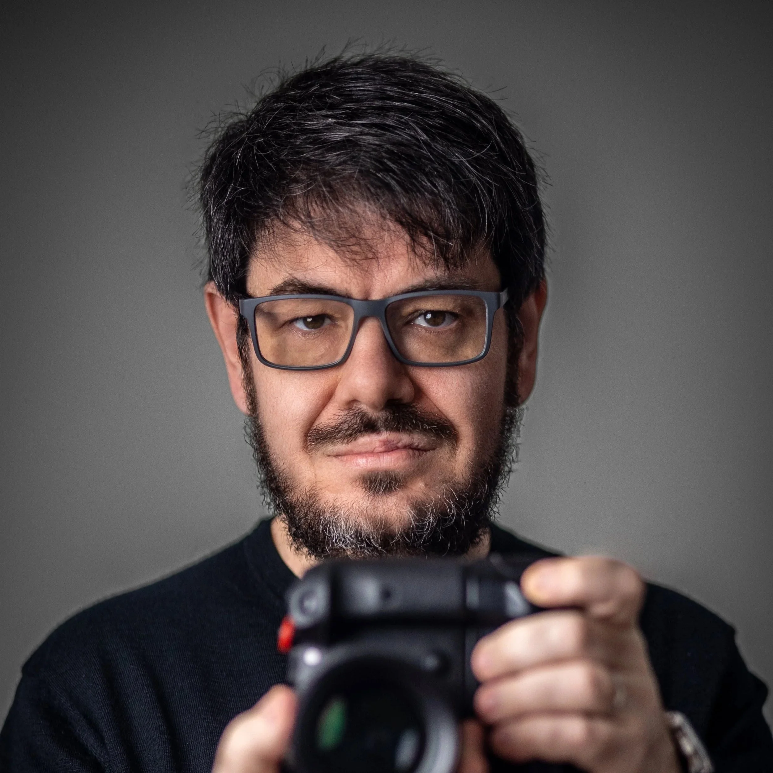 Alberto Pascual, professional photographer holding camera toward viewer in dramatic studio portrait with grey background.