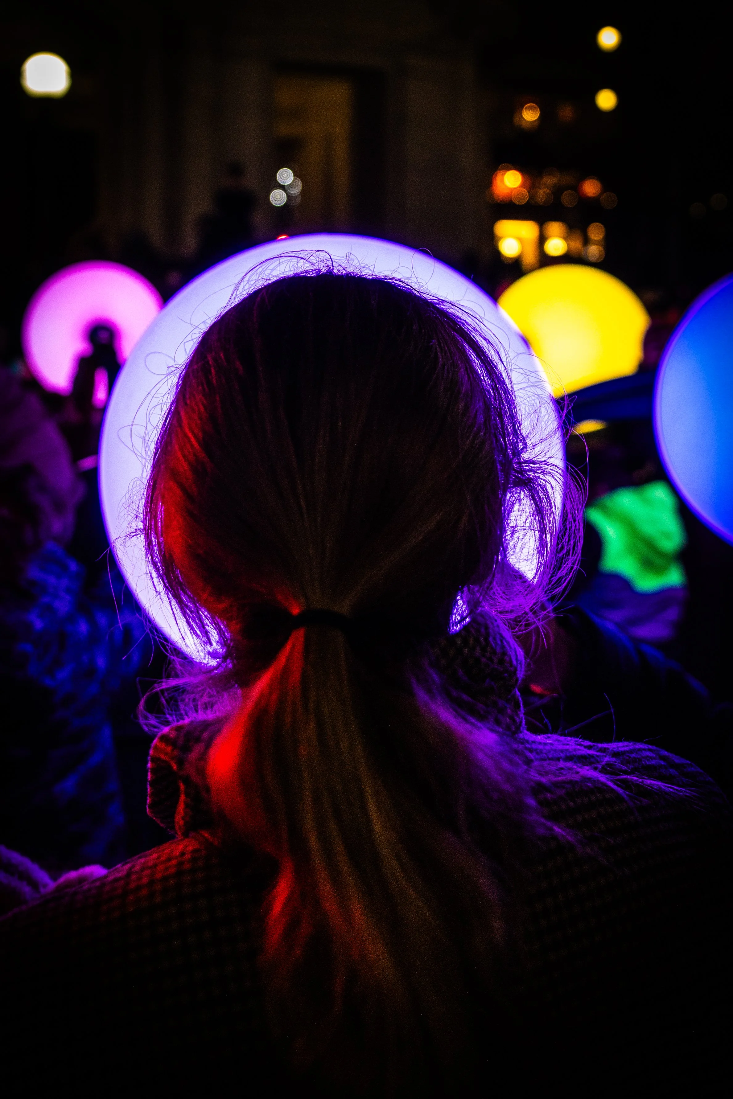 Night event photography of woman's silhouette with white light halo surrounded by multicoloured illuminated spheres at outdoor light festival.