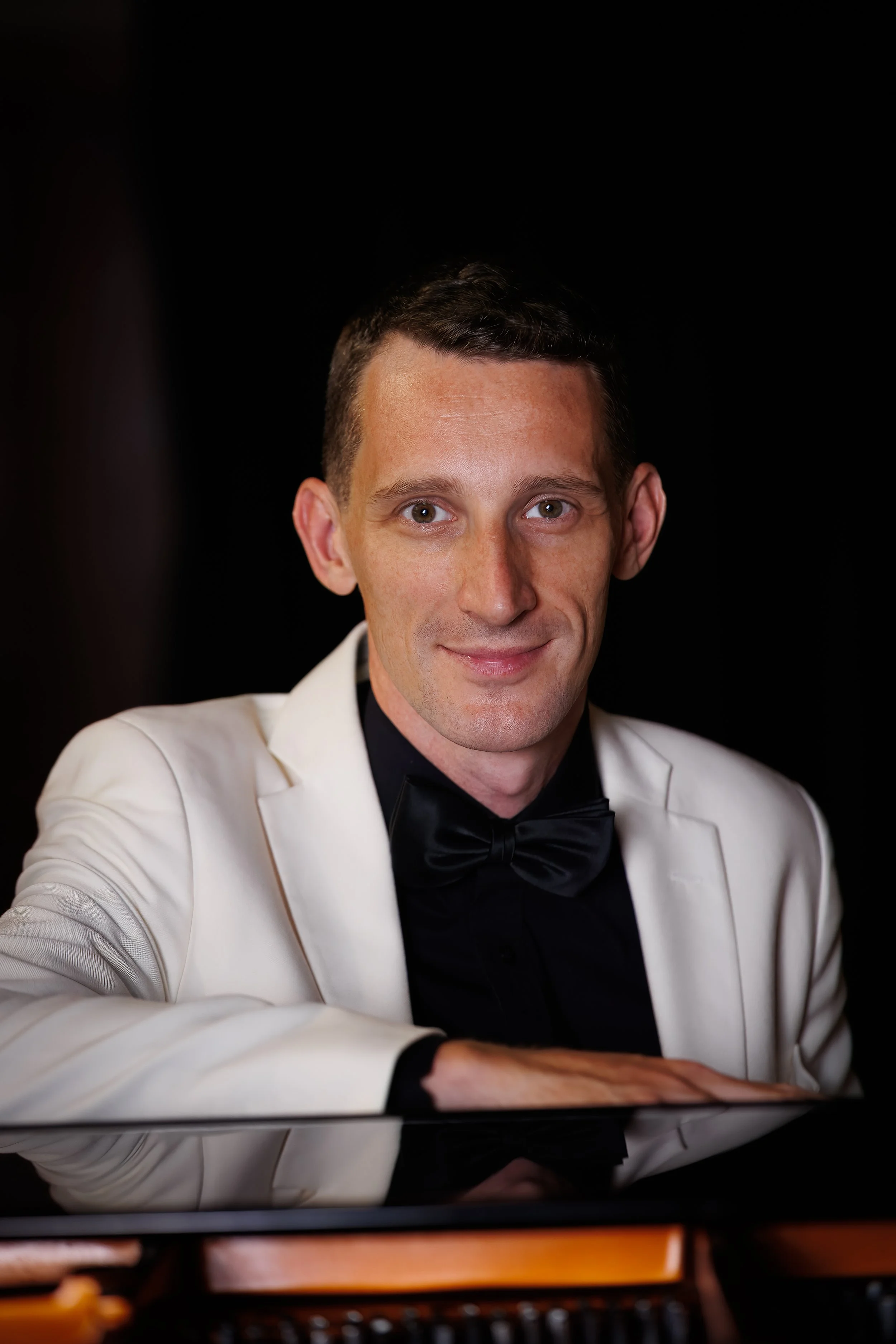 Professional portrait of male classical pianist in white tuxedo jacket and bow tie leaning over illuminated grand piano interior.