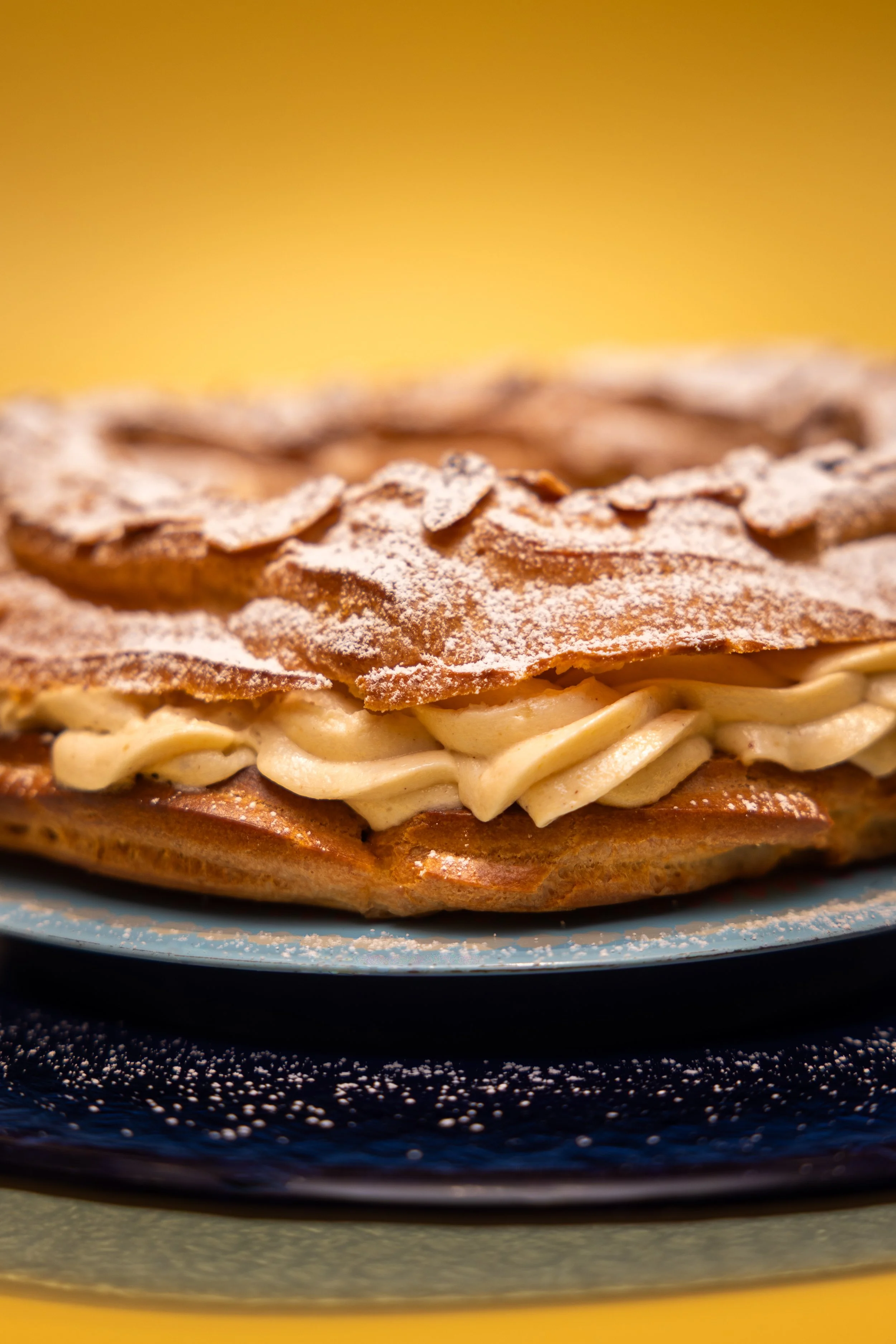Commercial food photography of Paris-Brest pastry with praline cream filling and toasted flaked almonds on blue ceramic plate with yellow background.