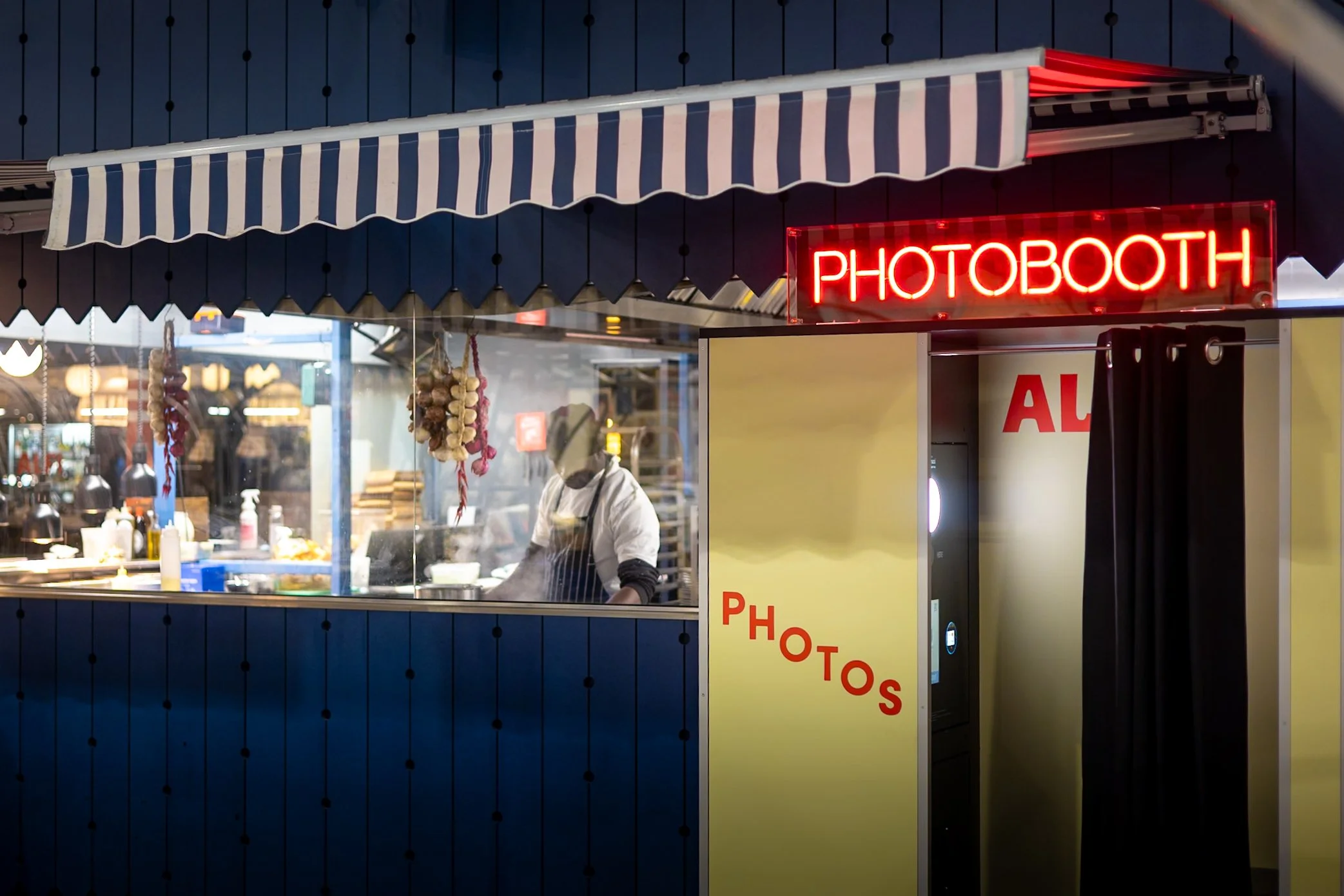 Night urban street photography of retro photobooth neon sign beside street food stall with chef and hanging garlic in indoor market.
