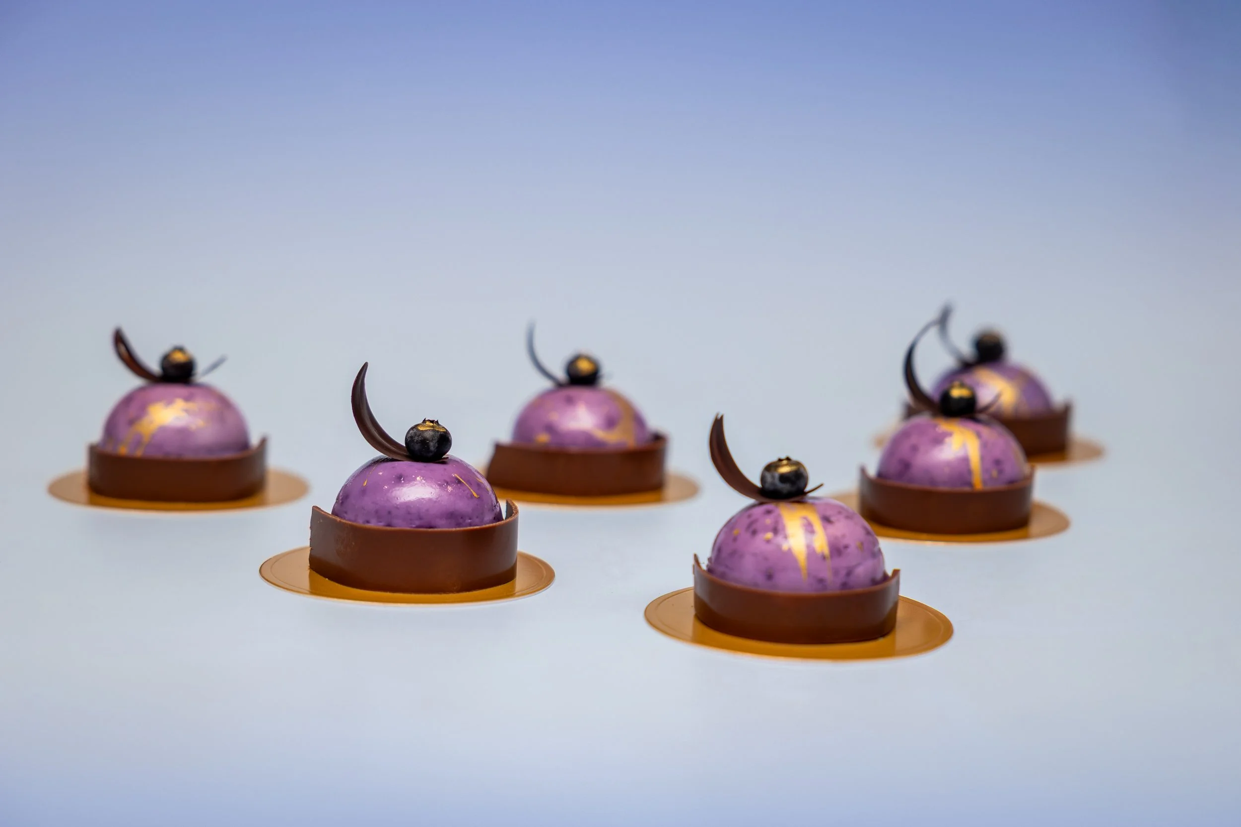 Commercial food photography of luxury purple iridescent chocolate dome desserts with gold detail and chocolate garnish on blue background.