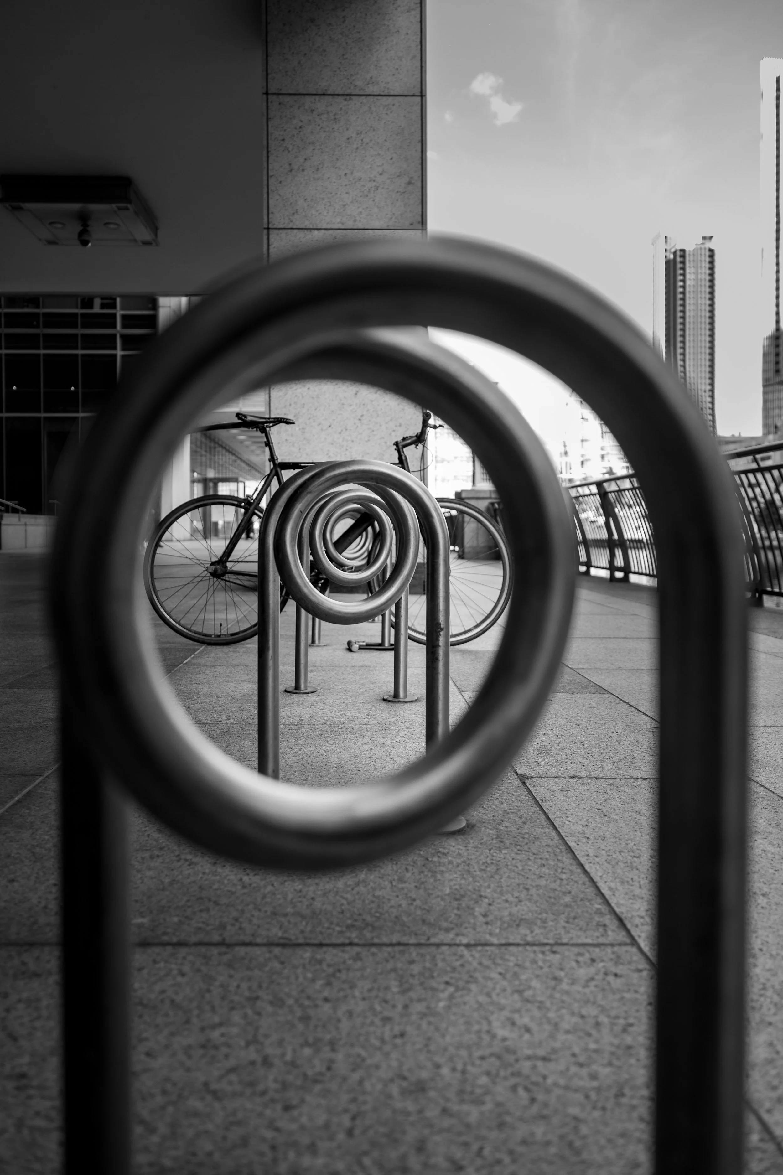 Black and white urban photography of bicycle and city skyline seen through spiral metal railing in creative street composition.