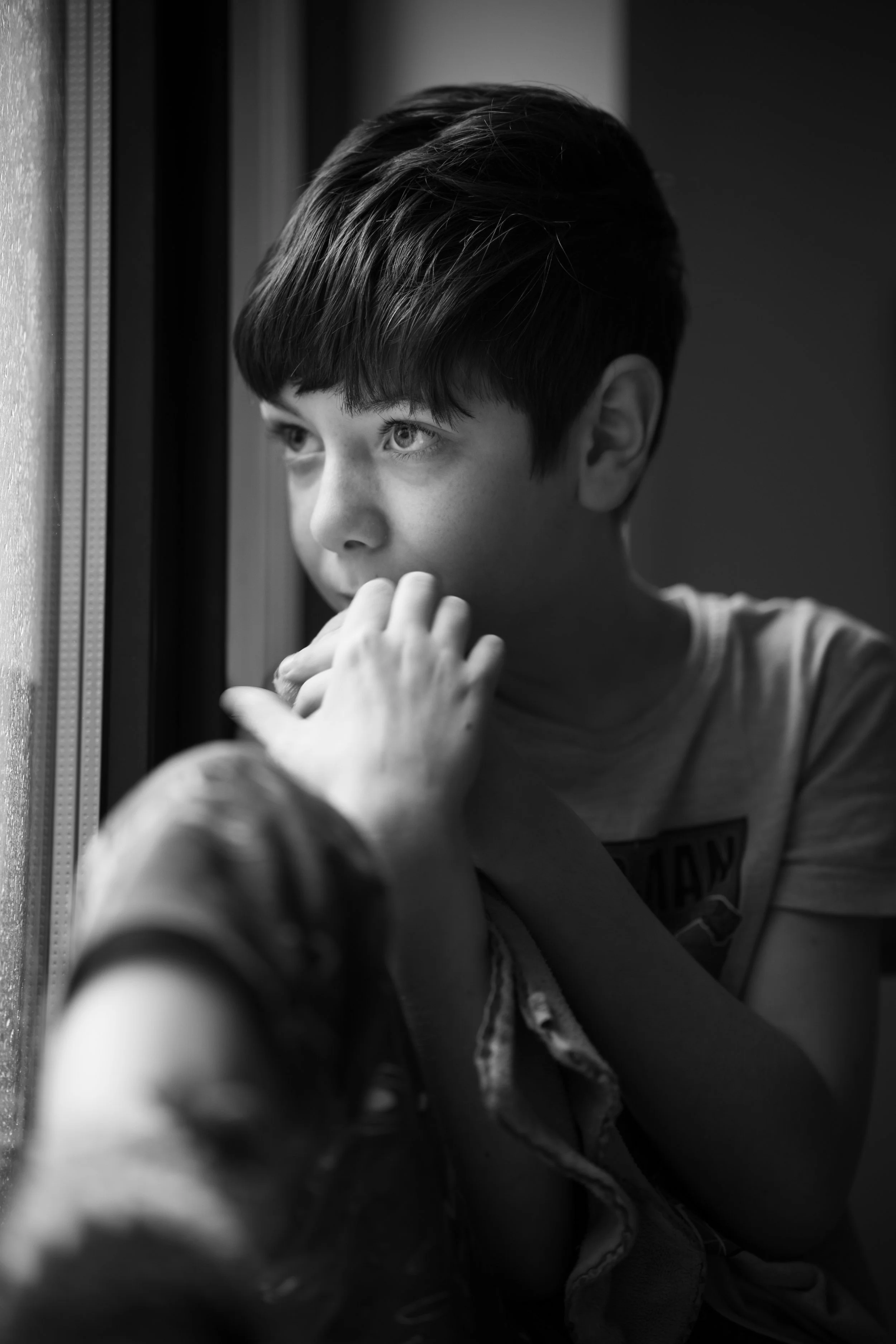 Fine art black and white childhood portrait of thoughtful boy gazing out window with natural light and shallow depth of field.