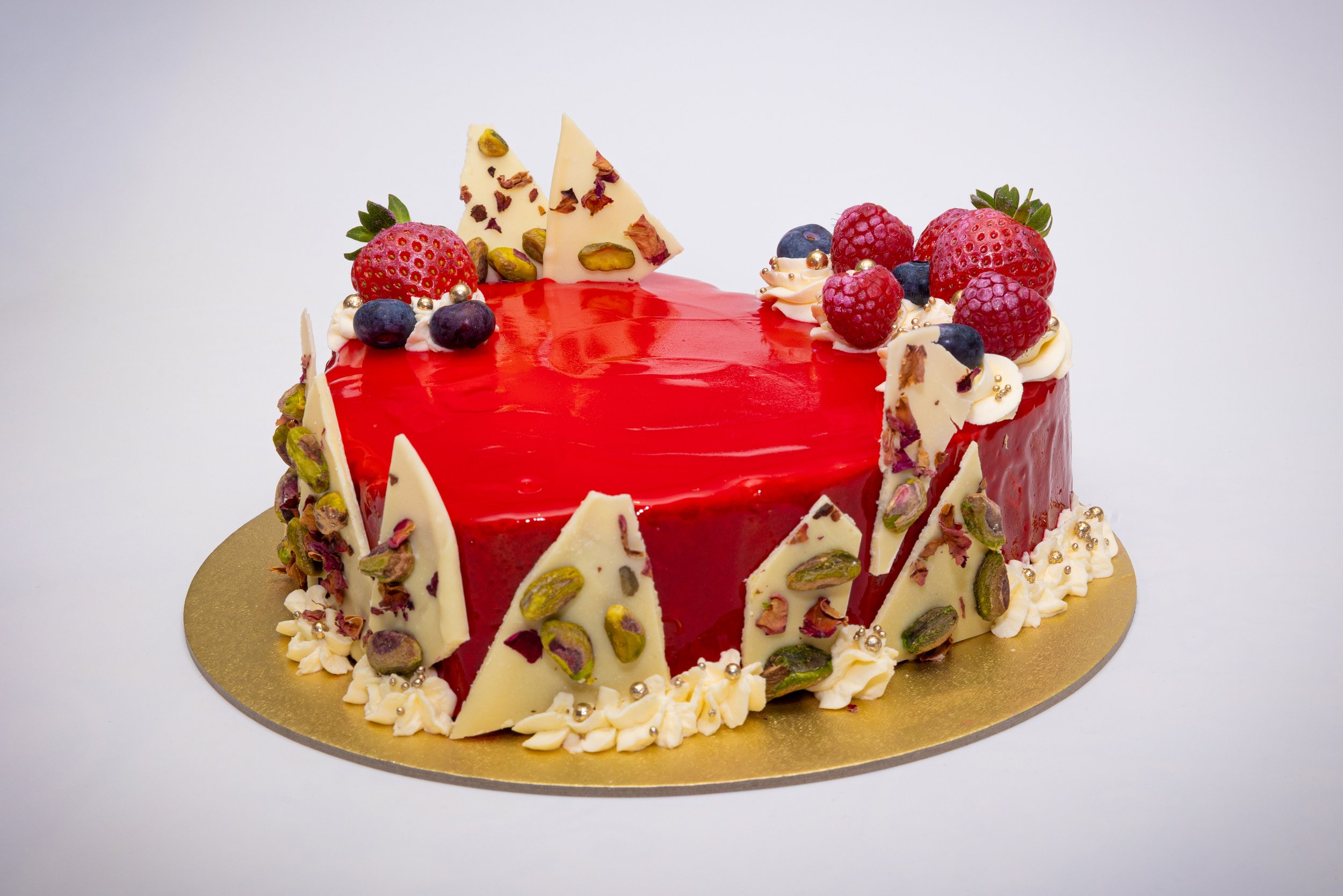Commercial food photography of luxury red mirror glaze cake with white chocolate pistachio bark, fresh berries and gold pearl details on white background.