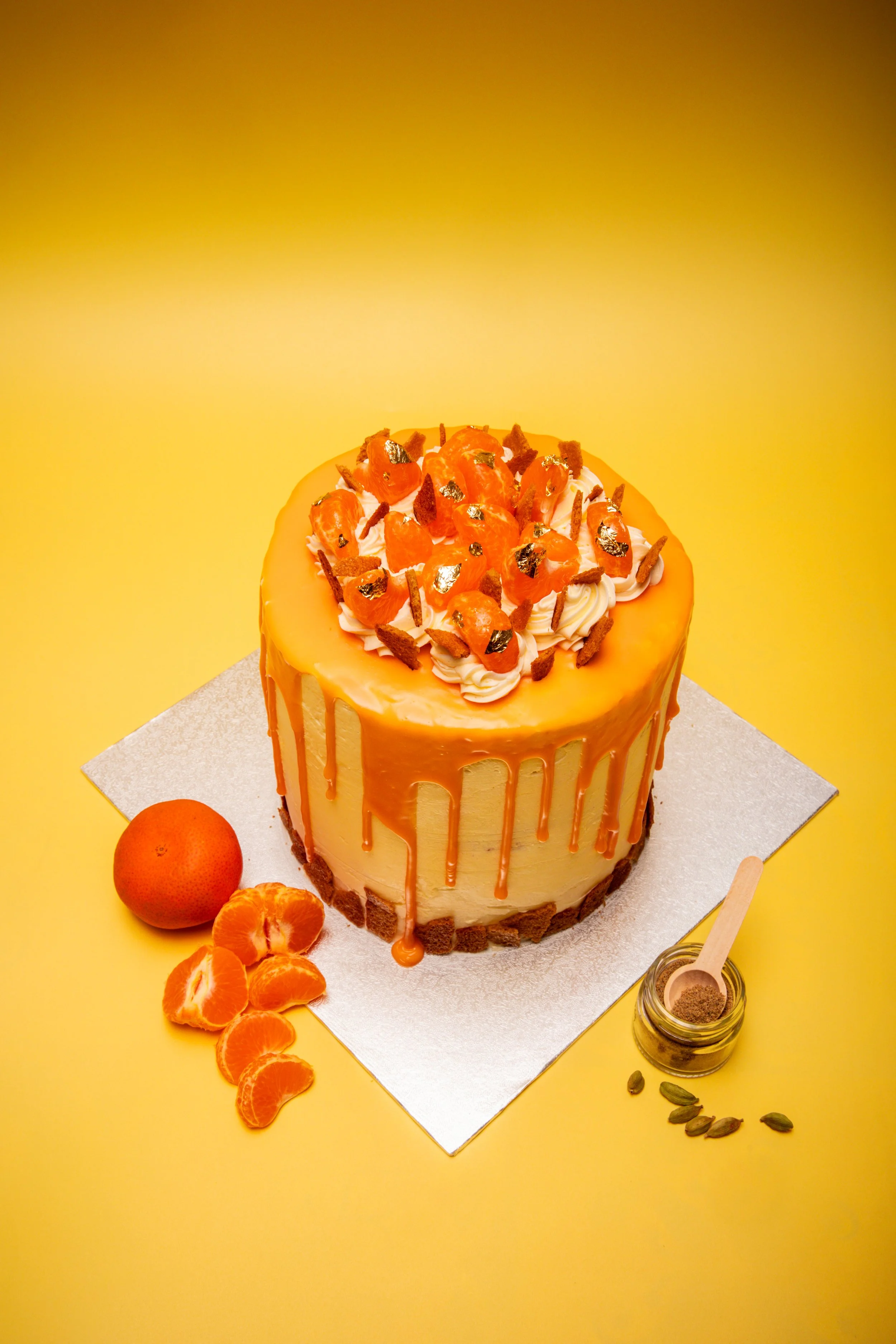 Commercial food photography of elegant caramel drip cake with gold leaf, mandarin oranges and cardamom styled on yellow background