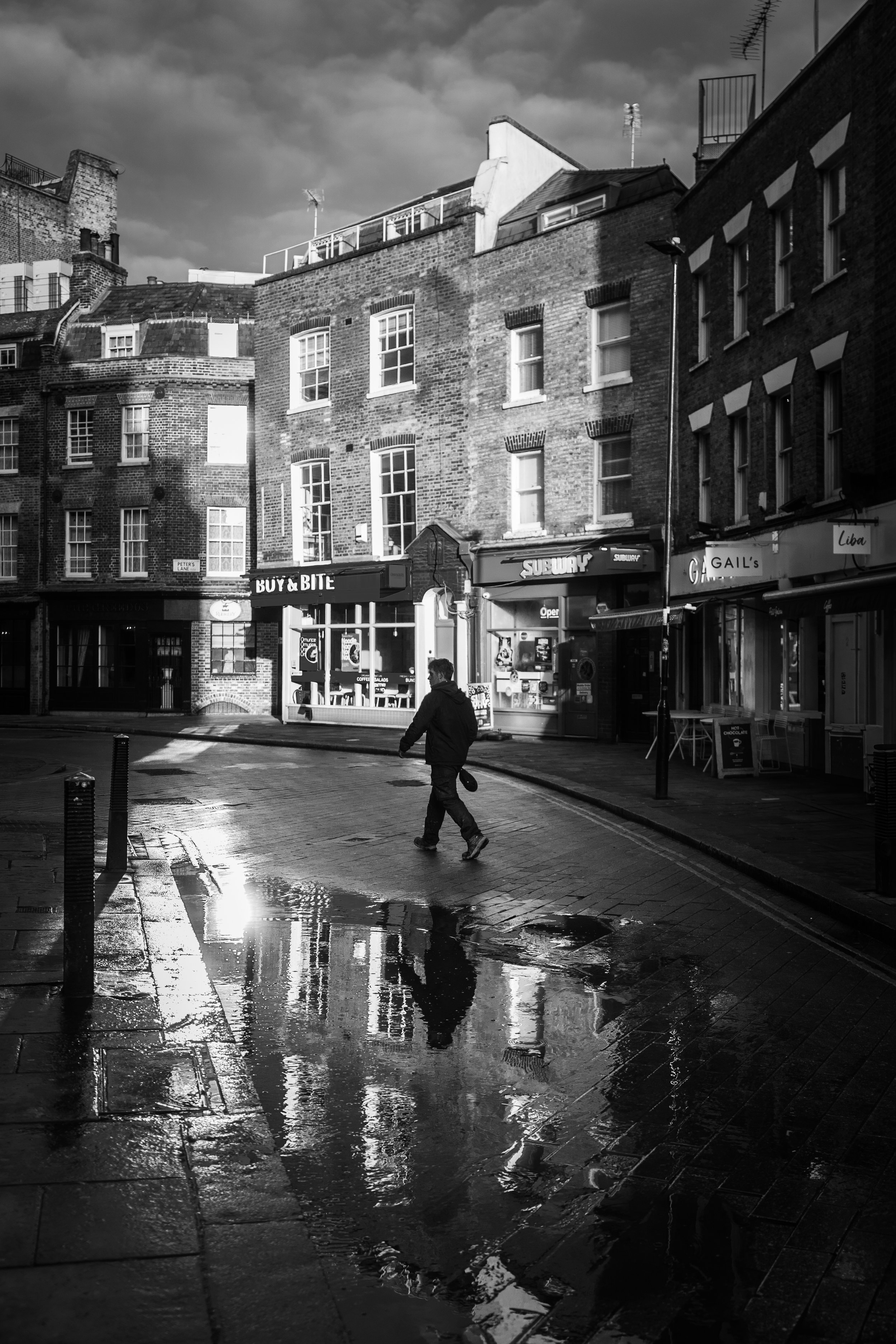 Black and white London street photography of lone figure crossing rain-soaked road with dramatic wet reflections and stormy sky over Georgian brick building.