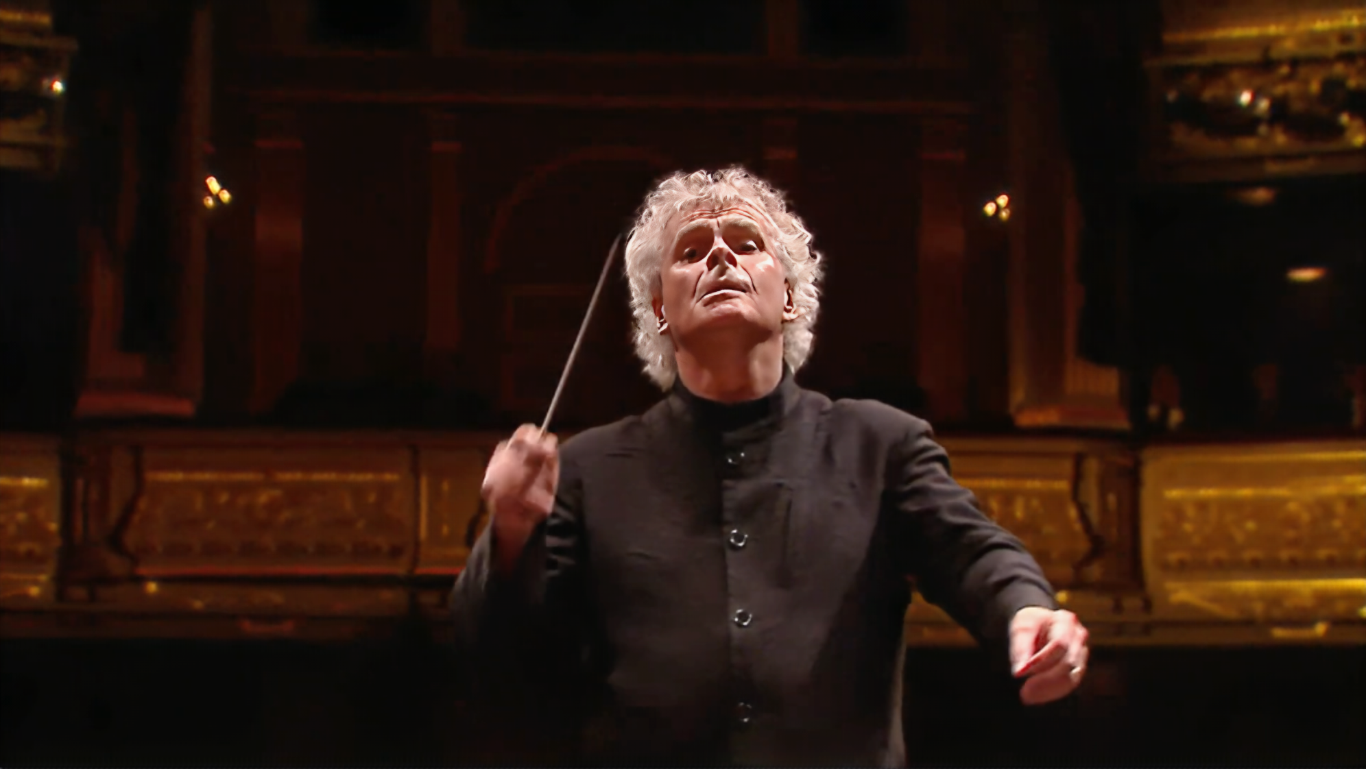 Film still of Sir Simon Rattle conducting  at Teatro Real Madrid with ornate golden auditorium backdrop.
