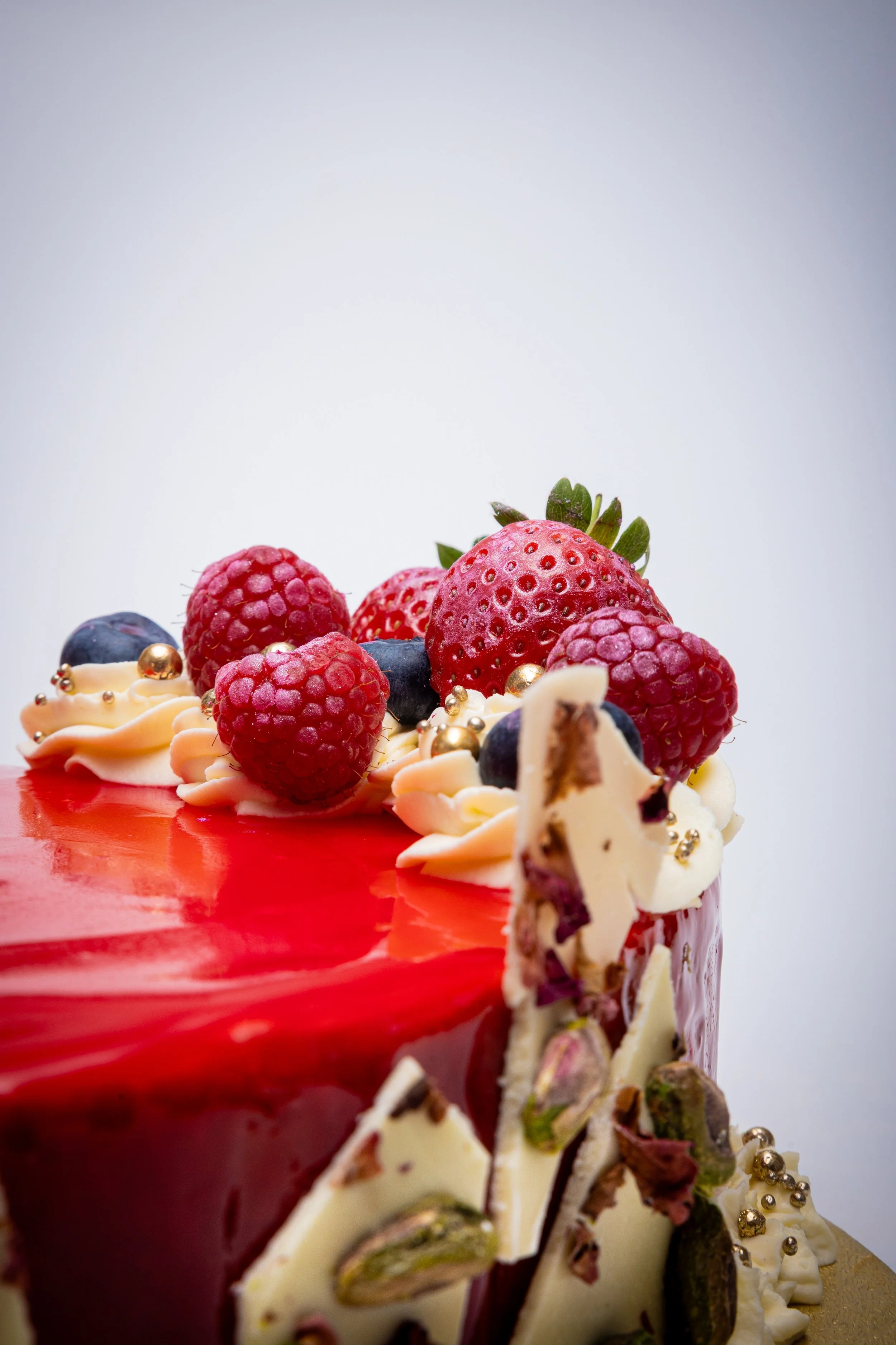 Luxury bakery food photography of mirror glaze celebration cake with fresh strawberries, raspberries, blueberries and gold pearl details.