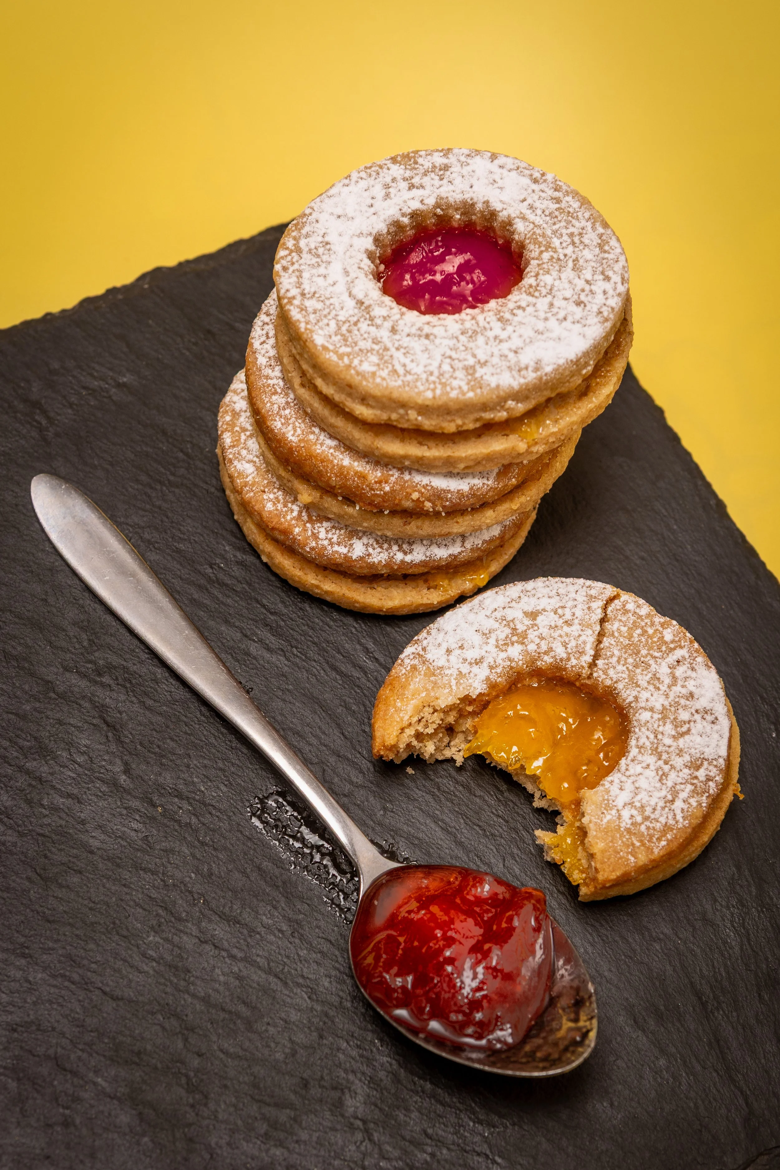 Professional styled food photography of stacked jam shortbread cookies with icing sugar on slate board, bitten cookie revealing apricot filling on a yellow background.