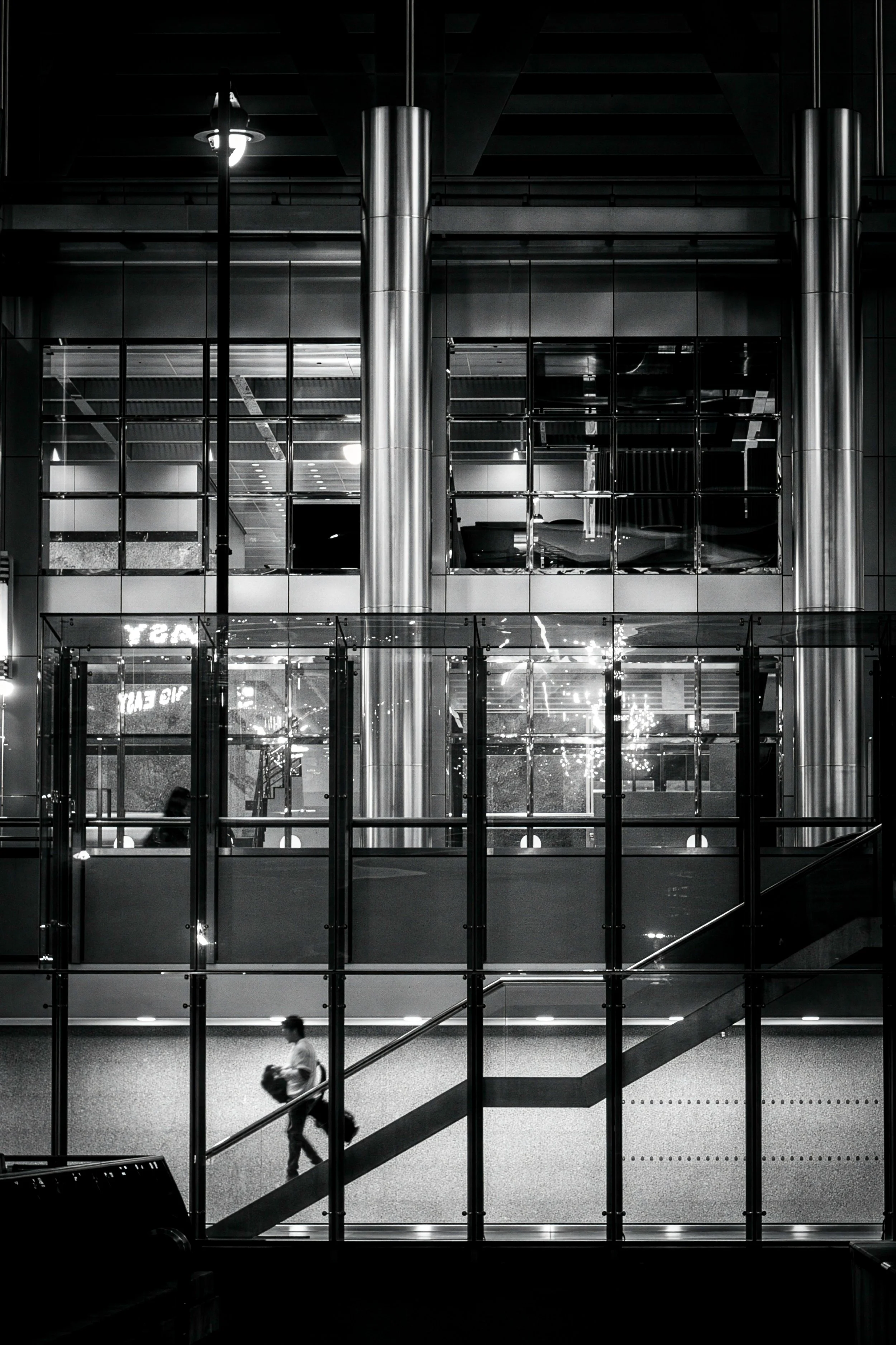 Black and white architectural urban photography of glass office building at night with lone figure on staircase and steel column reflections, Canary Wharf, London.