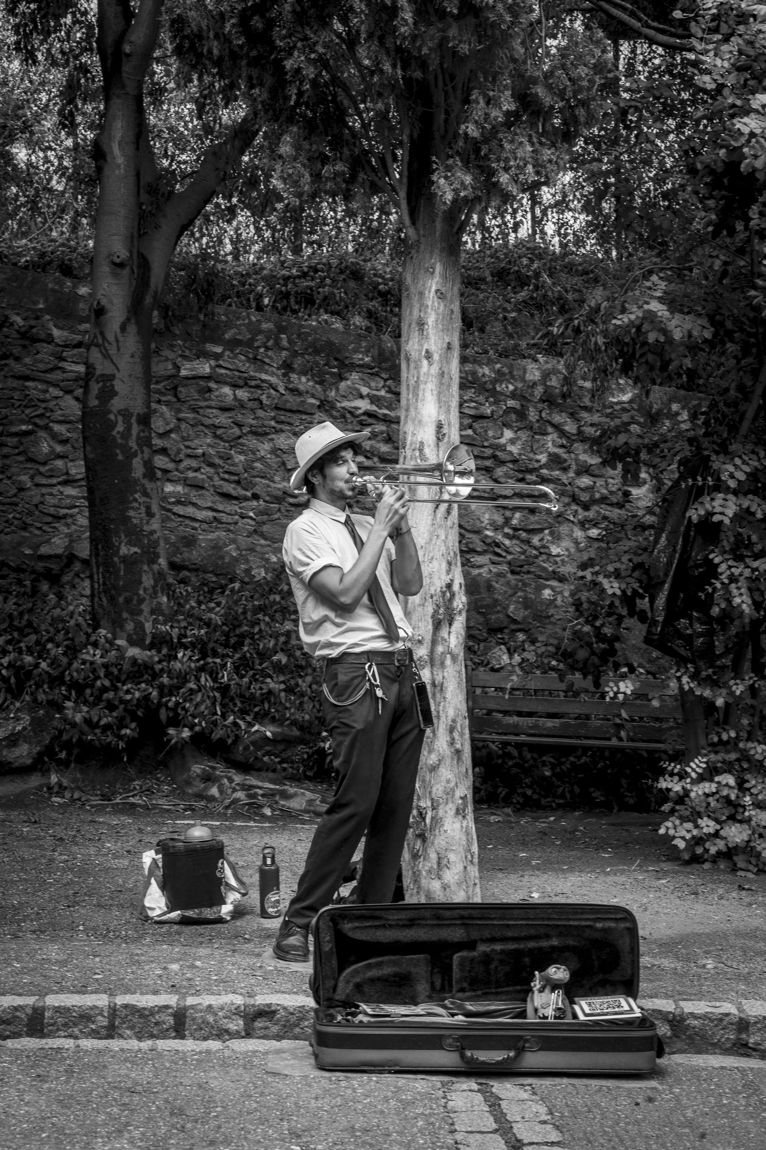Black and white street documentary photography of trombone busker performing in park with open case, cobblestone path and stone wall.
