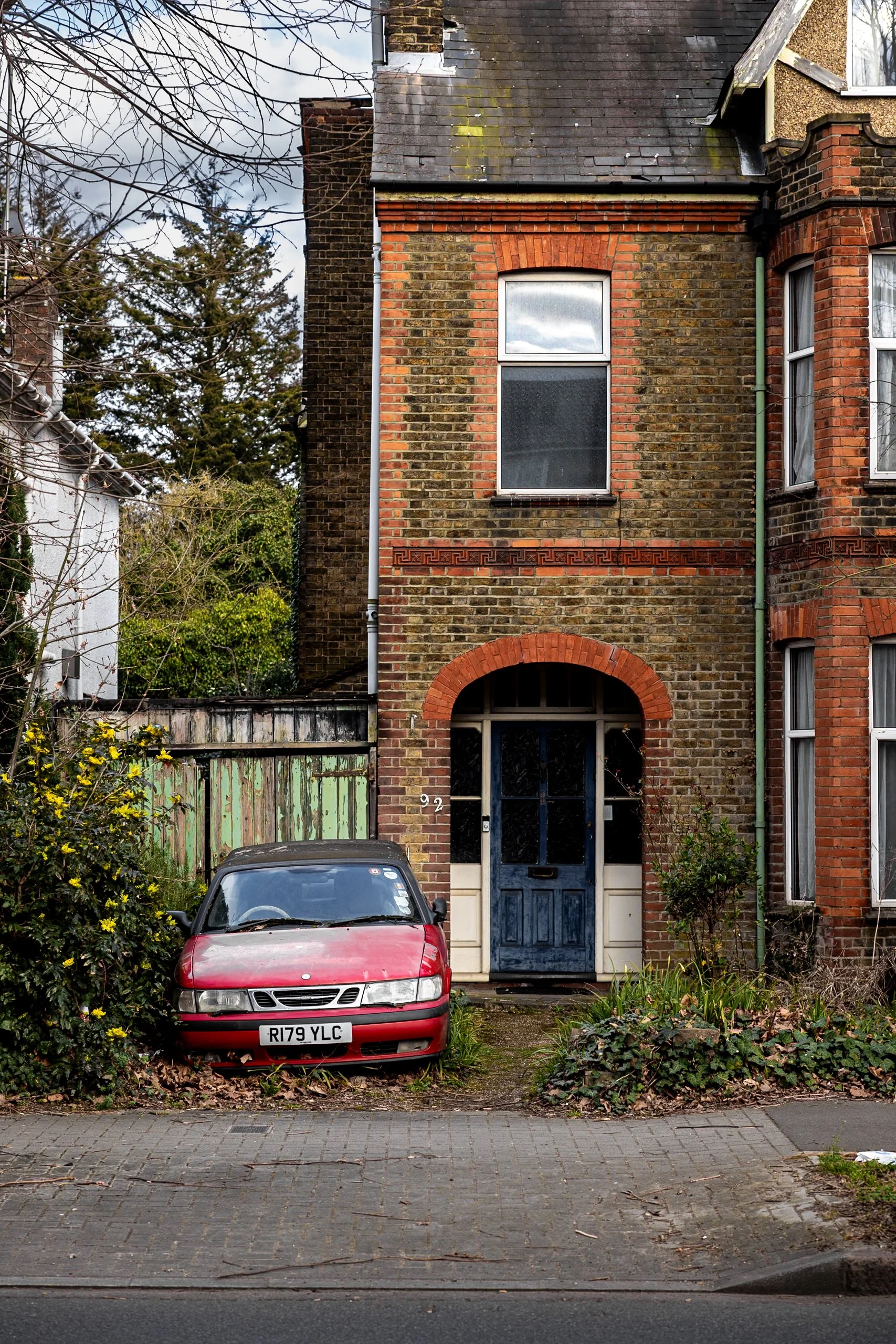 Street documentary photography of neglected Victorian terraced house with blue arched door and abandoned vintage red car in overgrown London garden.