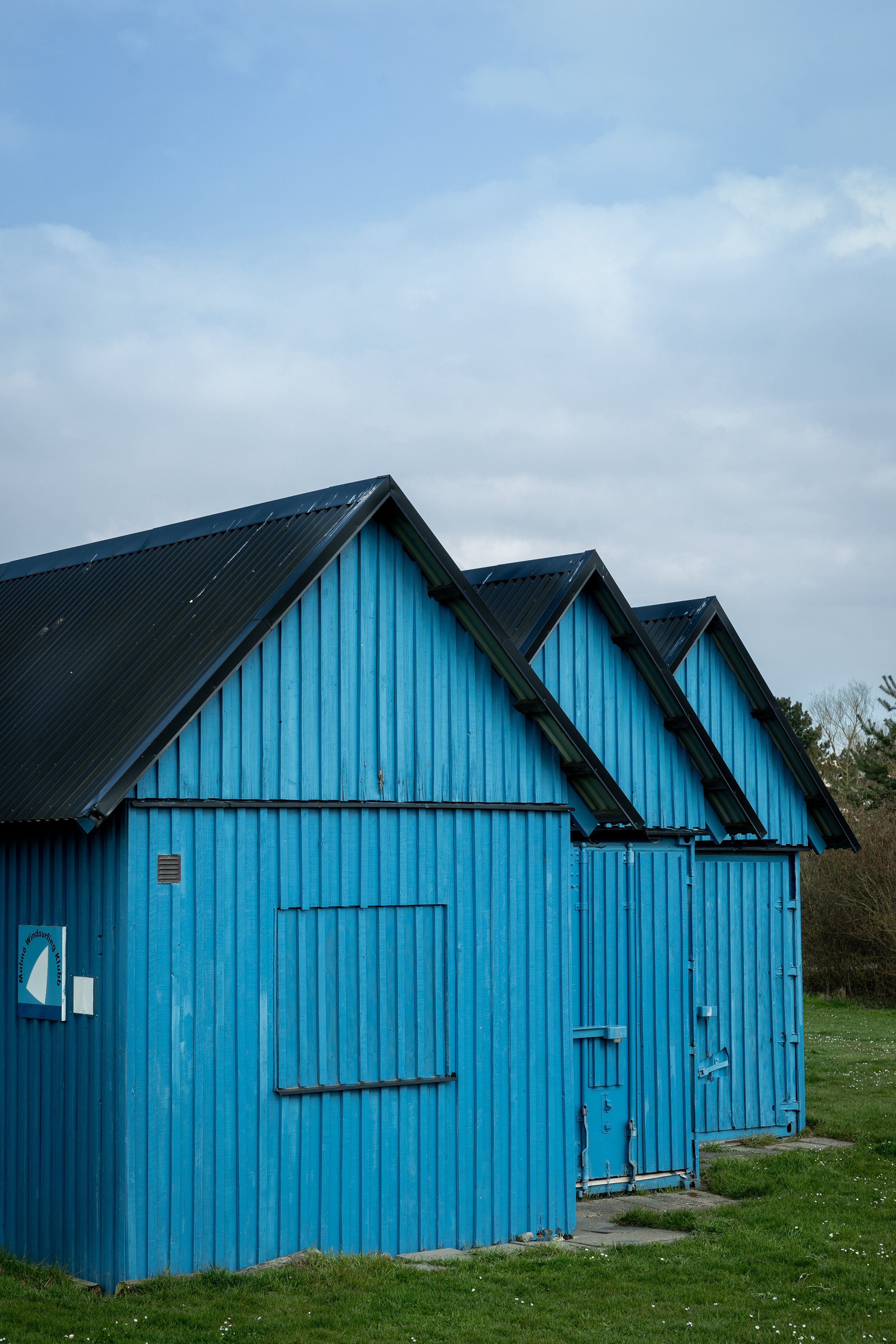 Landscape photography of row of bright blue wooden sheds with black corrugated roofs on green grass against cloudy sky.