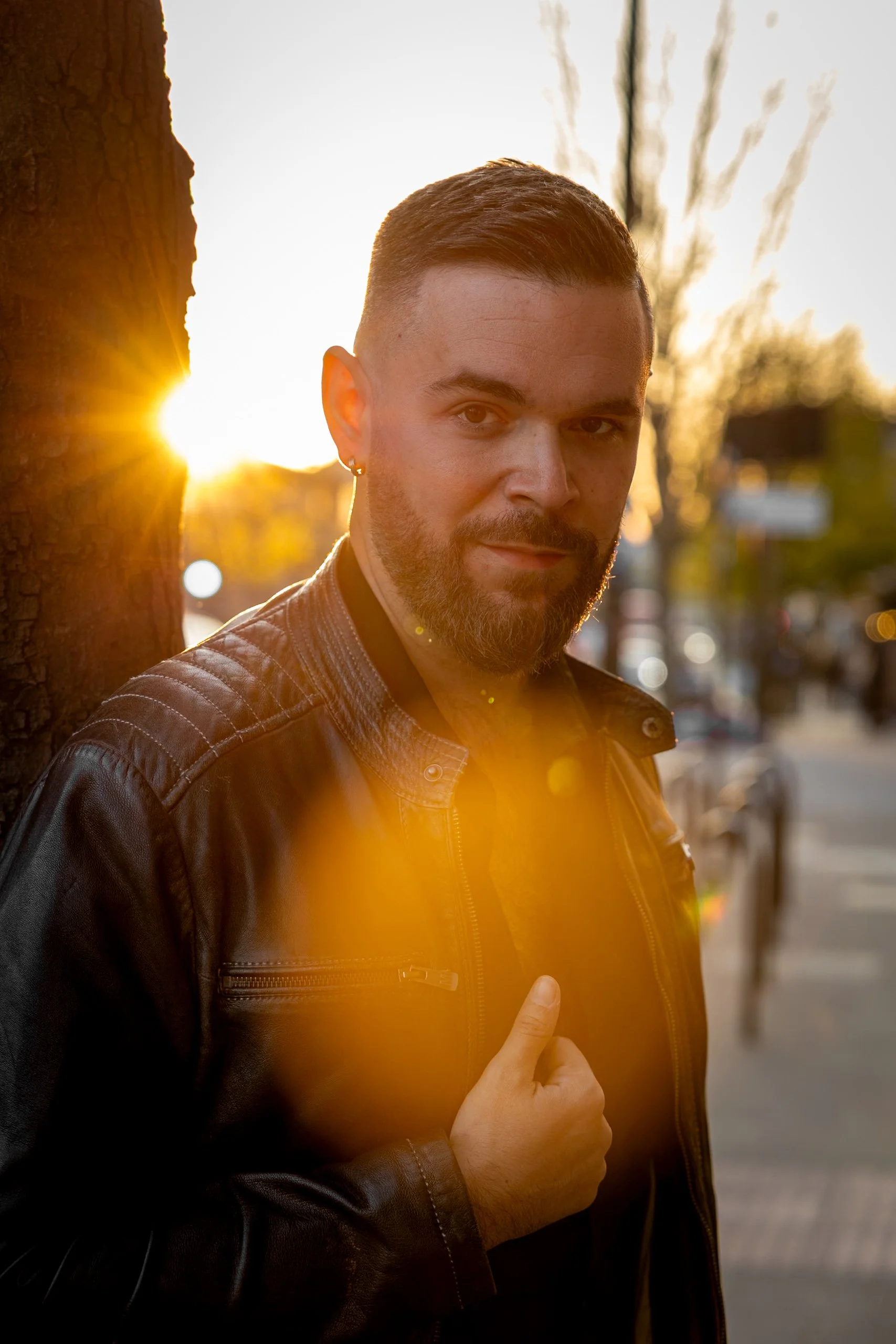 Professional golden hour outdoor portrait of man in leather jacket with dramatic sun flare beside tree on urban street.