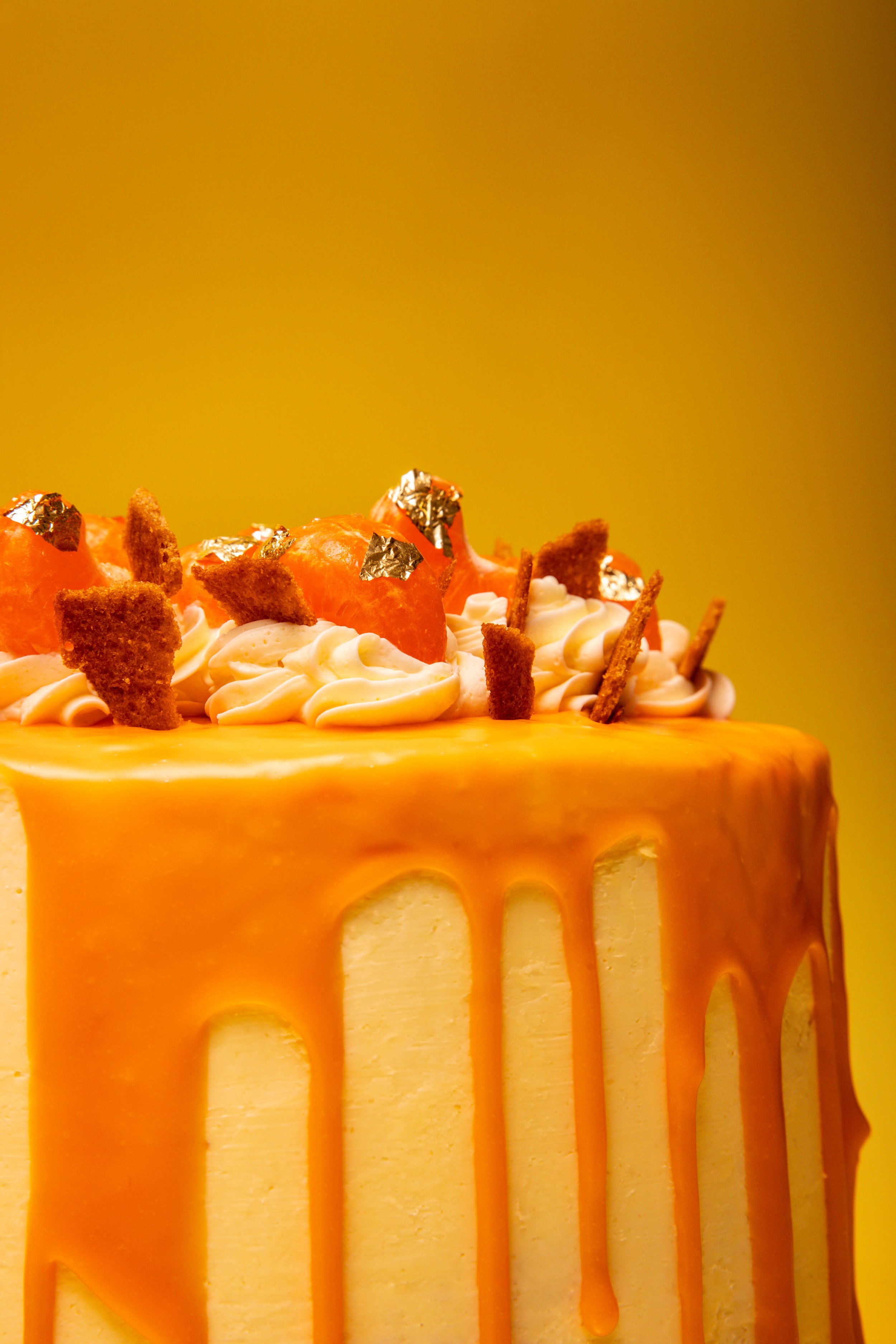Macro food photography of luxury caramel drip cake with gold leaf, cream rosettes and orange glaze against colour-matched yellow background.