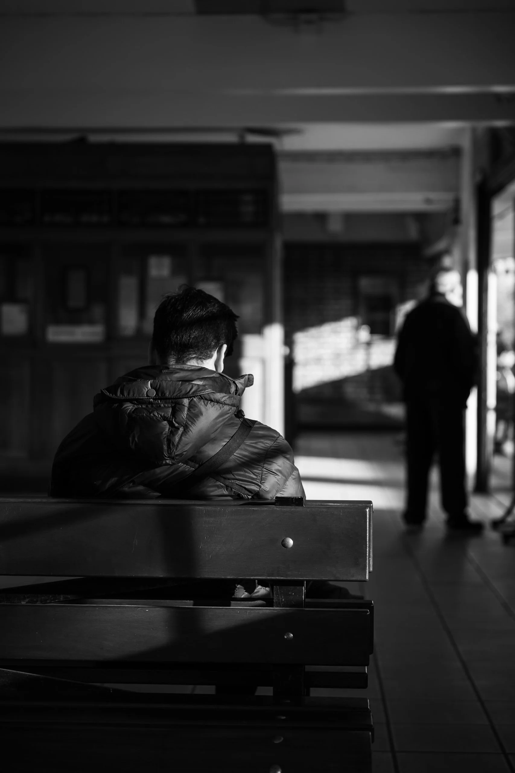 Atmospheric black and white documentary photograph of solitary seated figure on station bench with lone man silhouetted walking away in background.