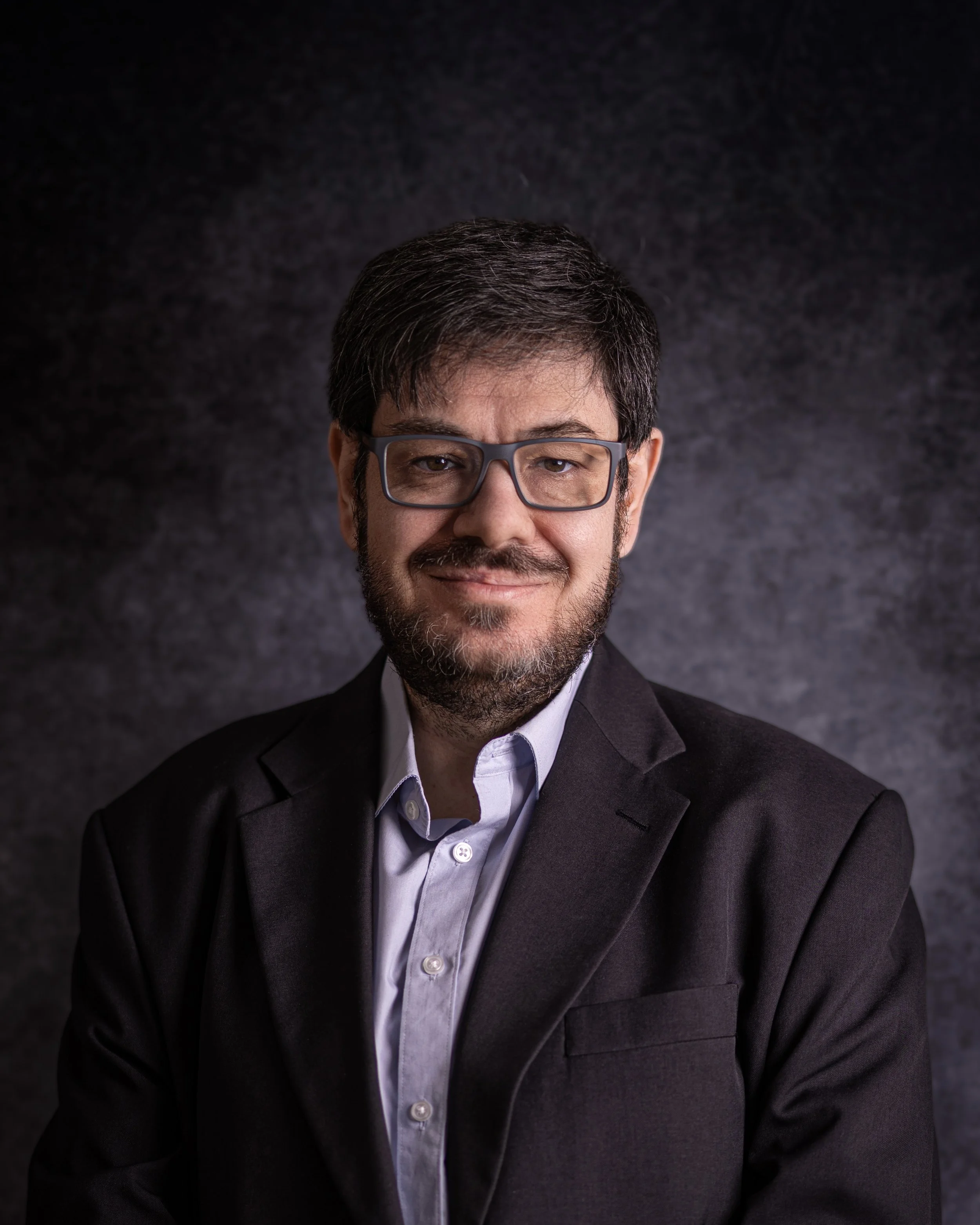 Close-up of Alberto Pascual, a man with dark hair, beard, and mustache, wearing glasses, a white shirt, and a dark blazer, smiling against a dark textured background.