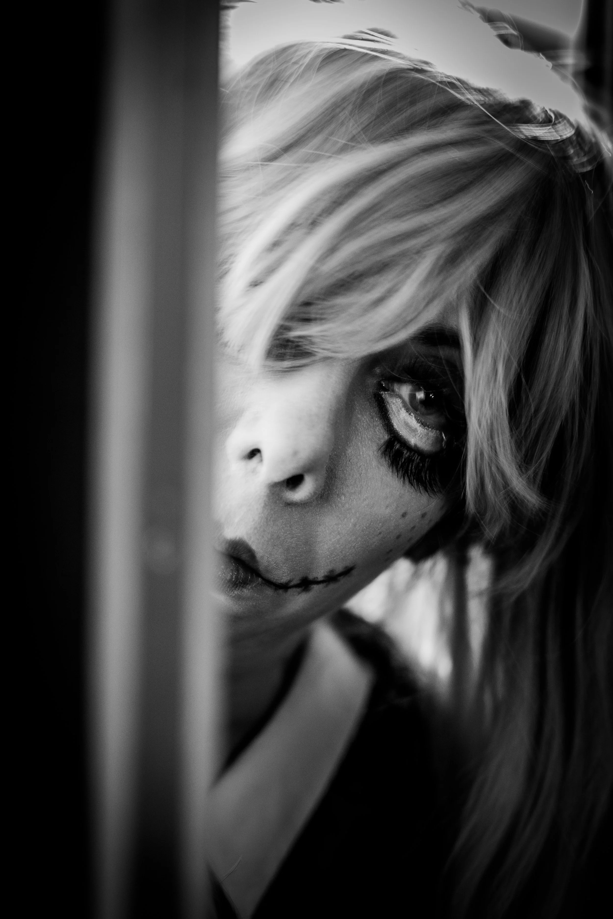 Cinematic black and white portrait of woman with dramatic theatrical makeup and stitched mouth effect peering through doorway.