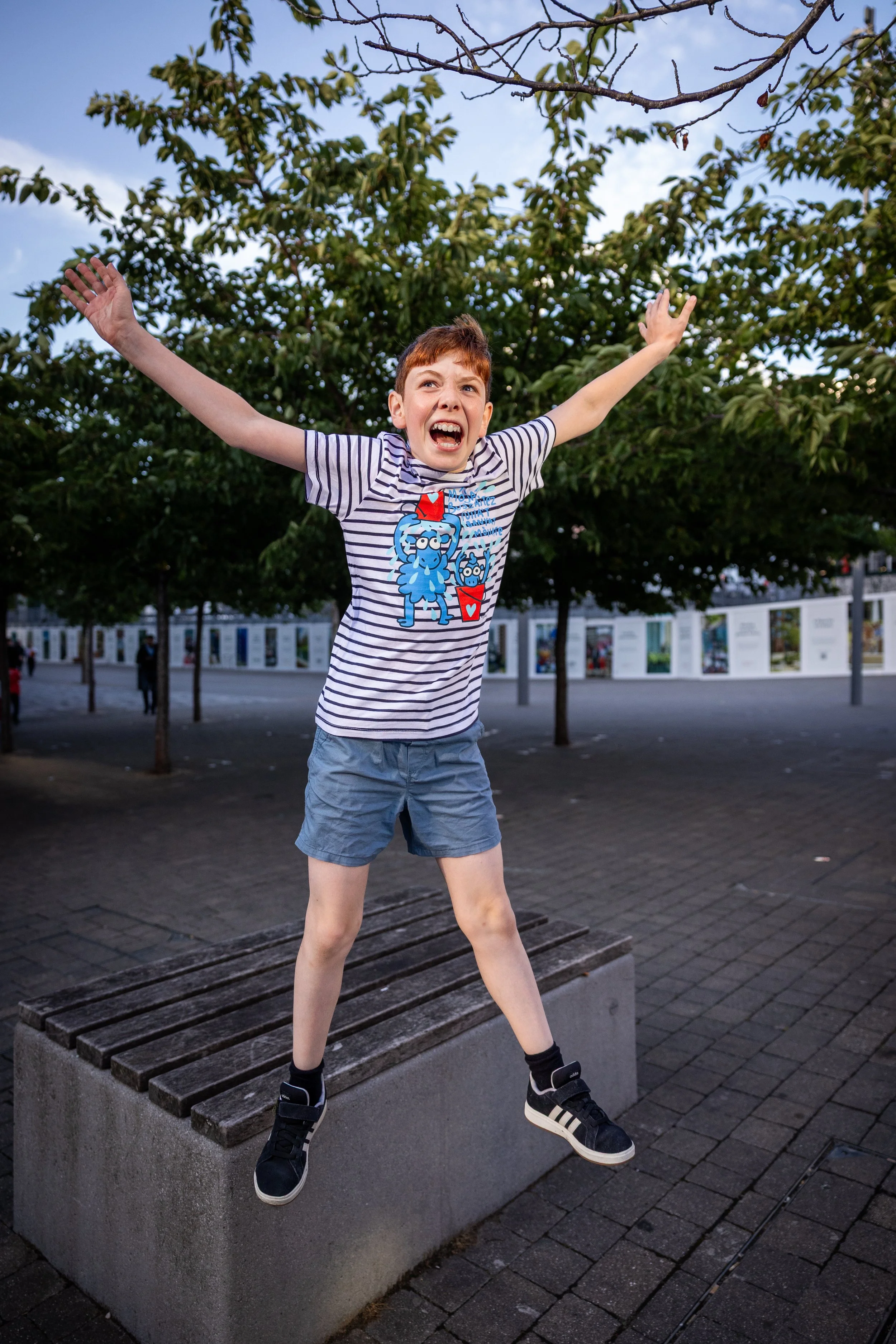 Professional outdoor children's portrait photography of joyful boy jumping from bench with arms outstretched in urban plaza.