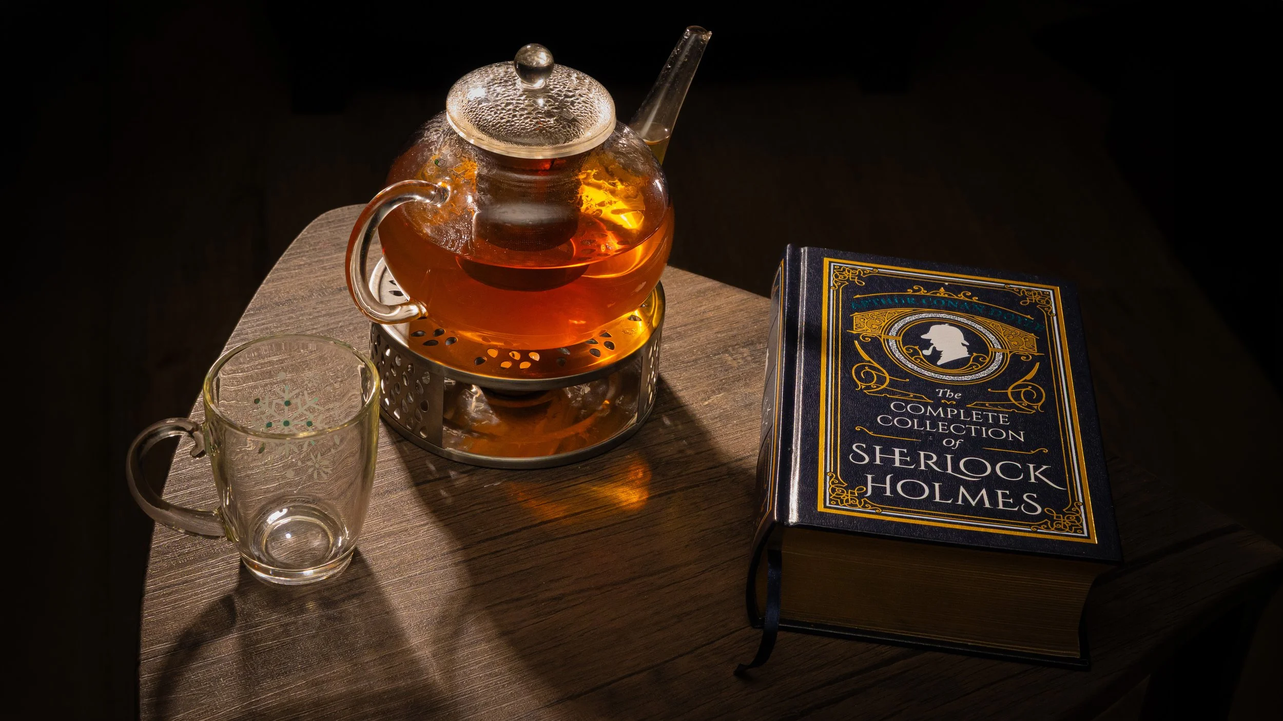 A glass teapot with tea, a clear glass mug with snowflake design, a hardcover book titled 'The Complete Collection of Sherlock Holmes', on a wooden table with a dark background