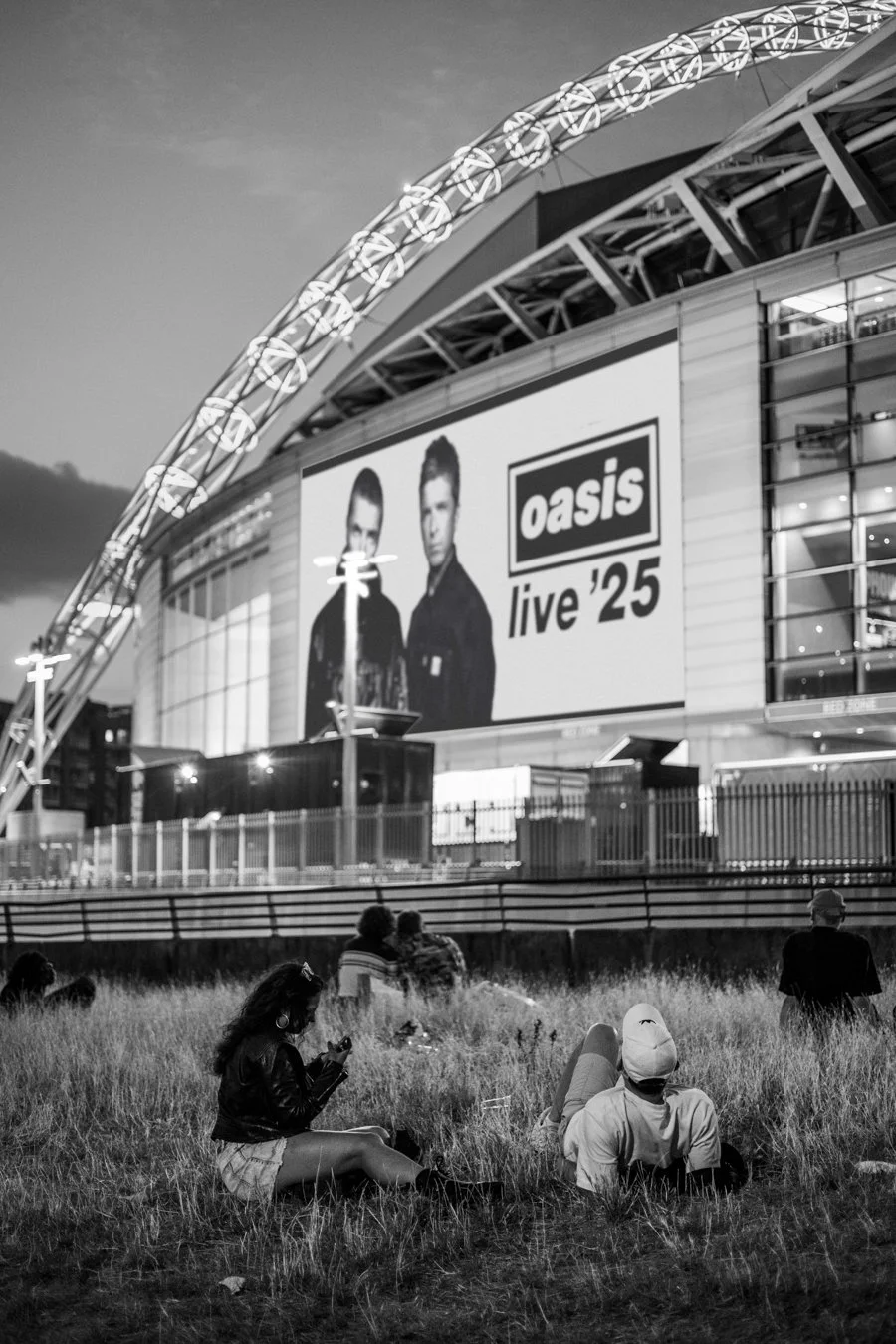 Documentary street photography of fans on grass outside Wembley Stadium with Oasis Live 25 concert billboard at dusk in black and white.