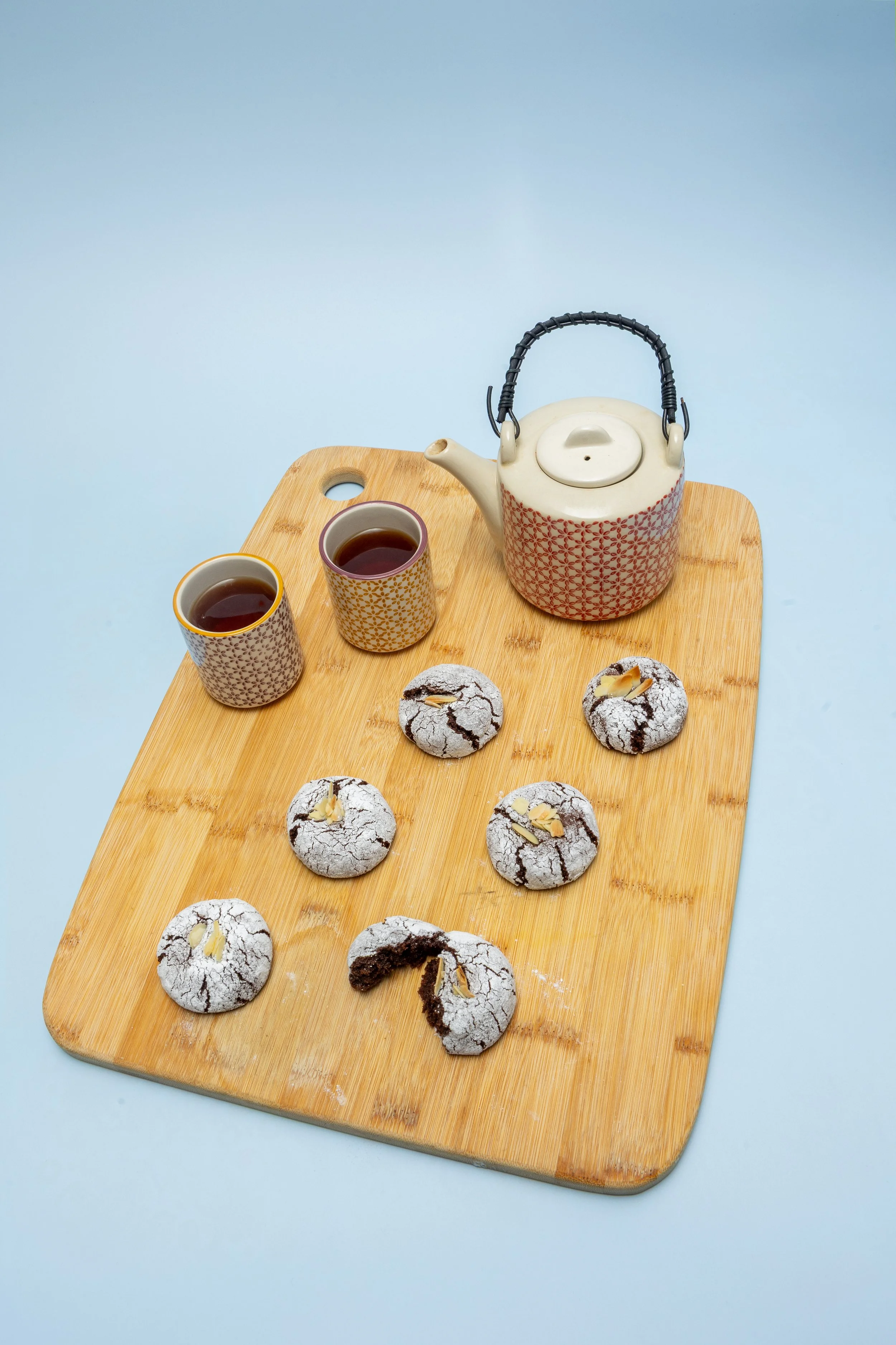 Professional food photography of chocolate crinkle cookies with almond garnish on bamboo board styled with ceramic tea set on pale blue background.