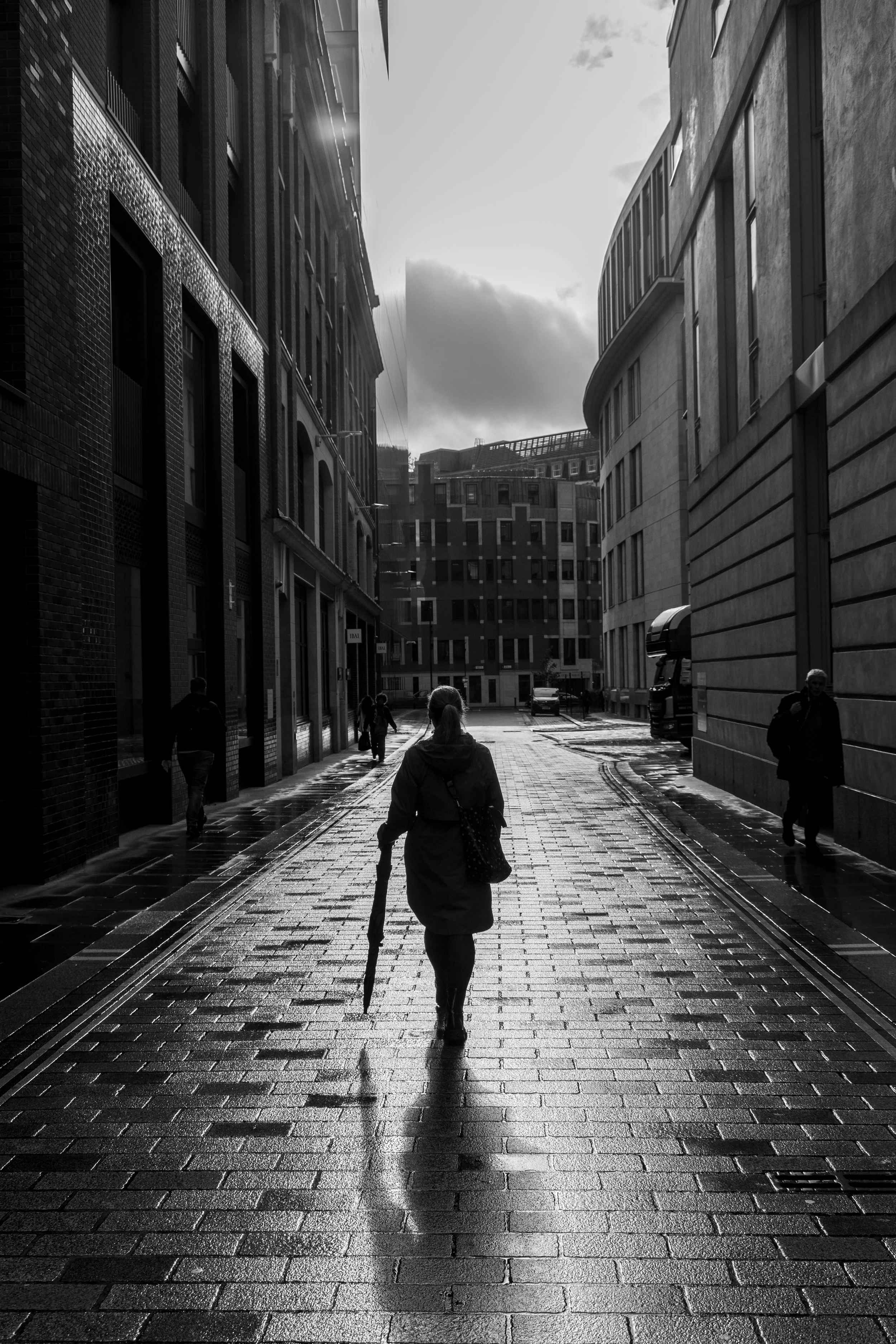Black and white London street photography of lone woman with umbrella walking wet cobblestone alley with dramatic contre-jour backlighting.