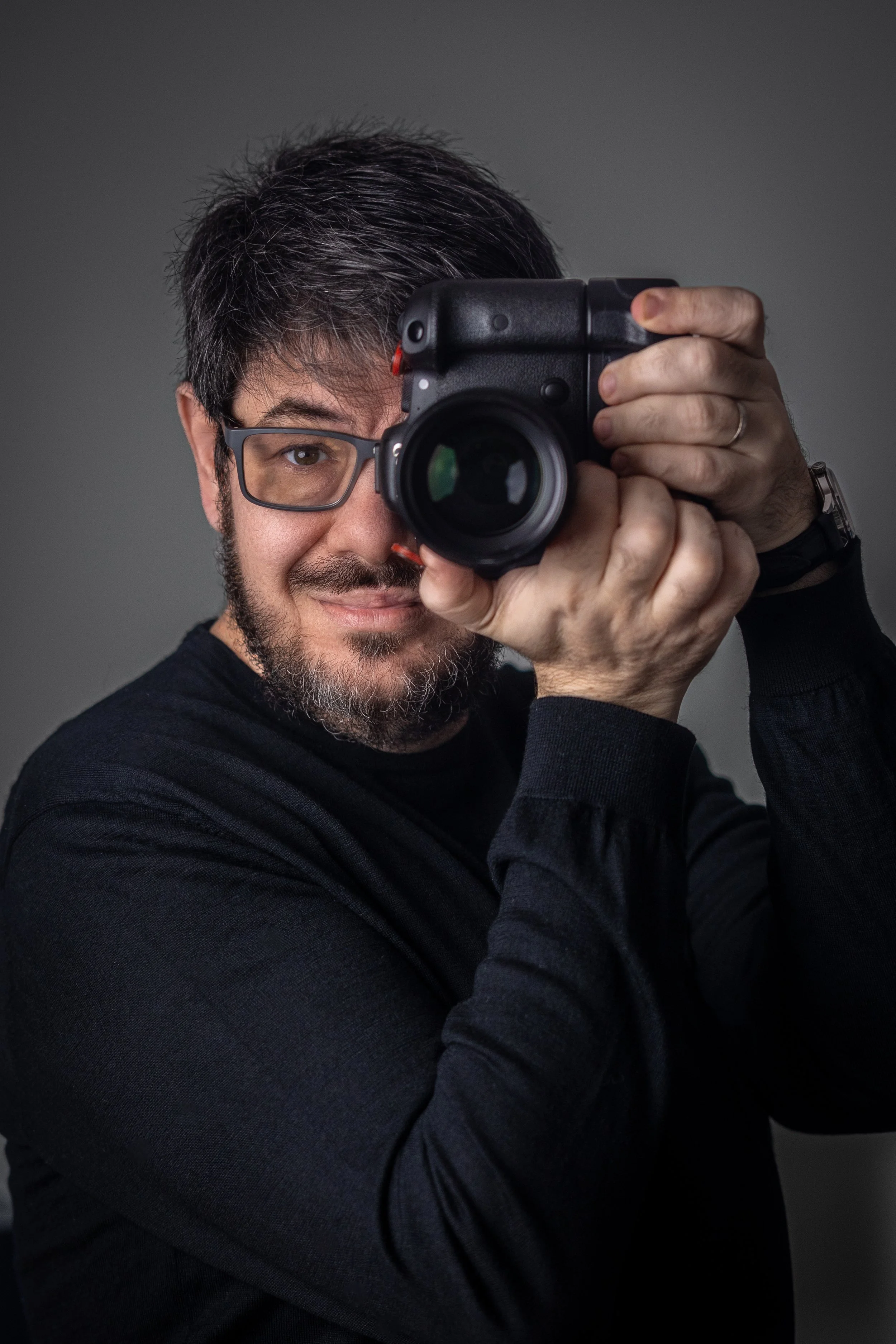 Alberto Pascual, professional photographer raising camera to eye level while smiling in dramatic studio portrait.
