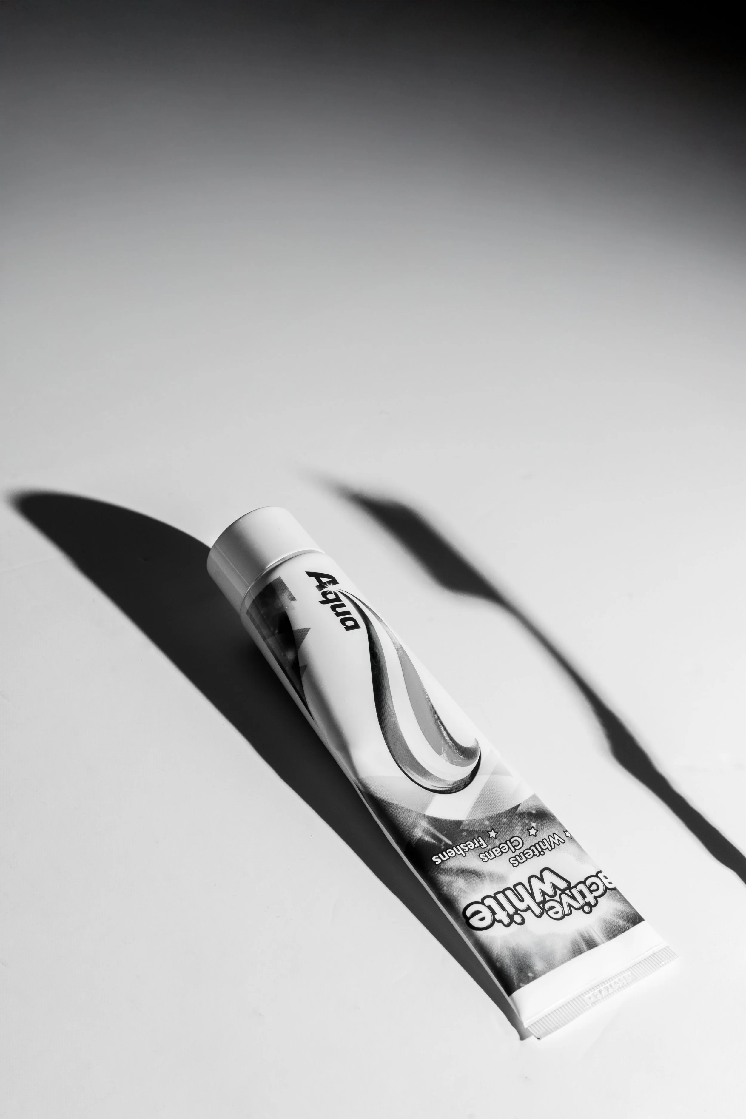 Cinematic black and white fine art photograph of a toothpaste tube casting elongated dramatic shadow paired with the shadow of a toothbrush, studio still life.