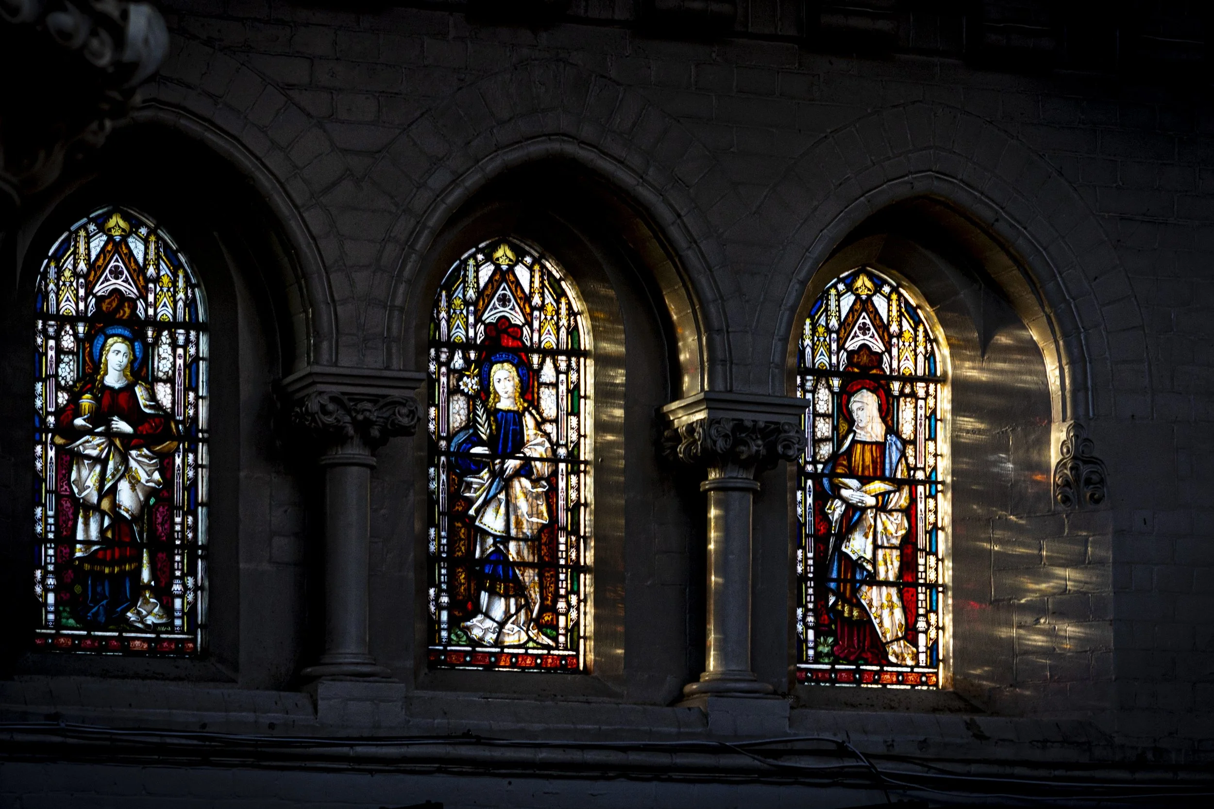 Interior architectural photography of three illuminated Gothic stained glass windows with religious figures in dark stone church interior.