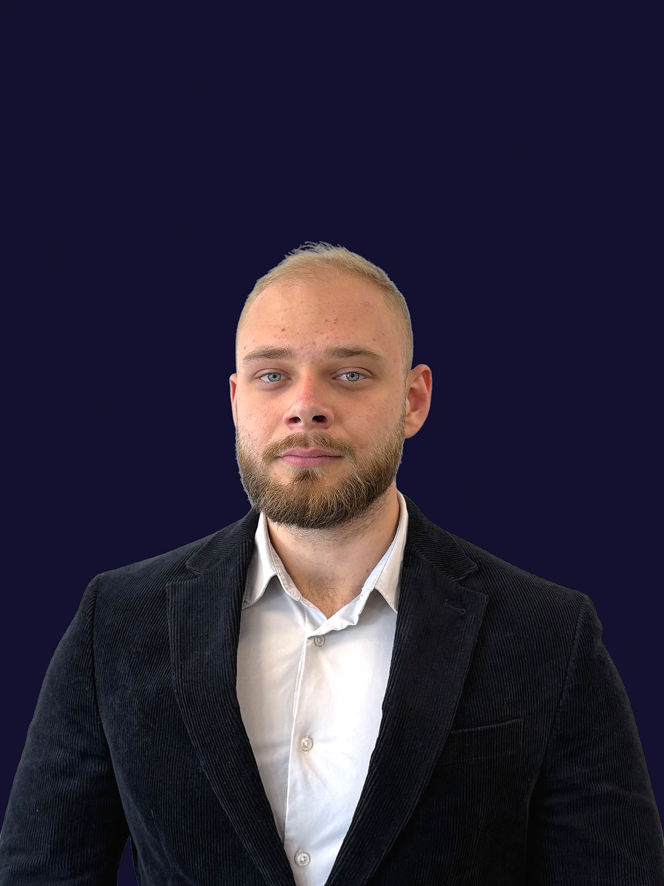 A man with a beard and short blond hair wearing a black blazer and white button-up shirt against a dark blue background. Martin Mach