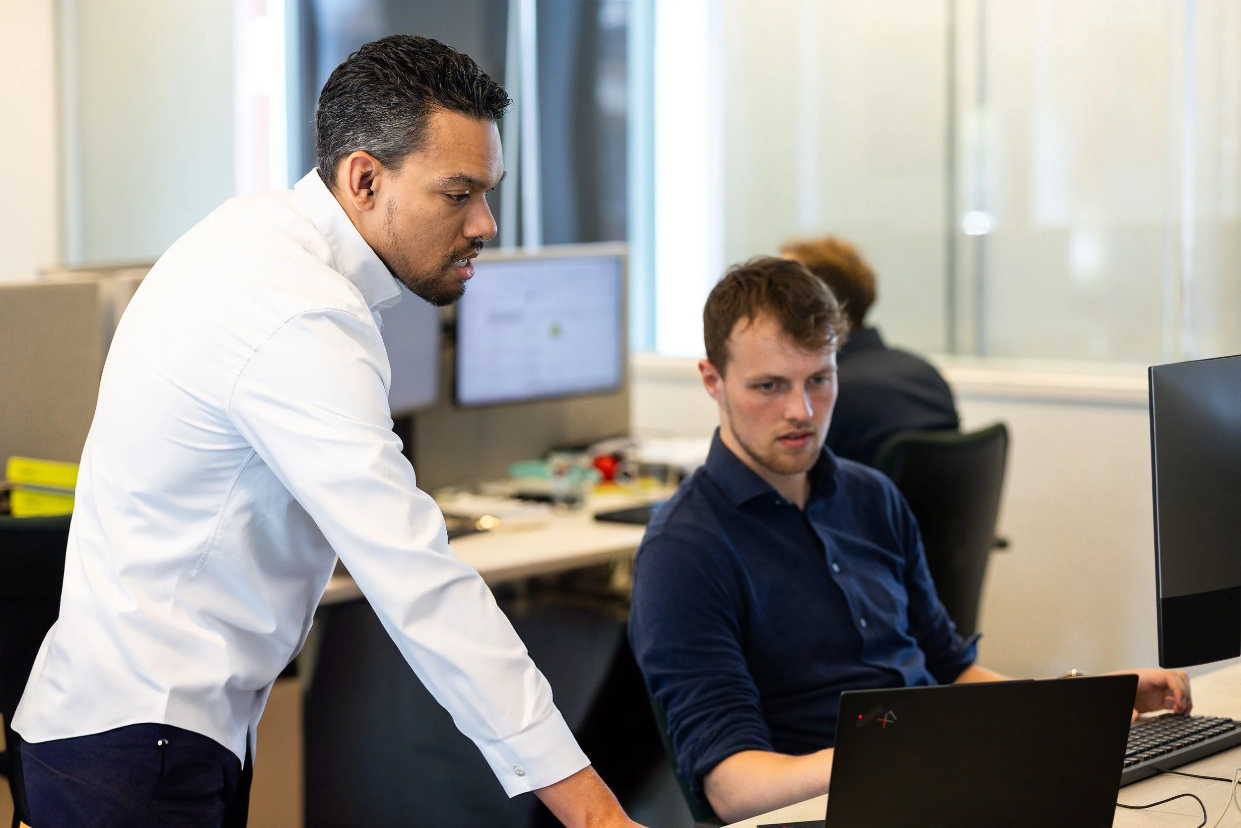 Two men working at desks in an office, one standing and the other sitting, both focused on computer screens.