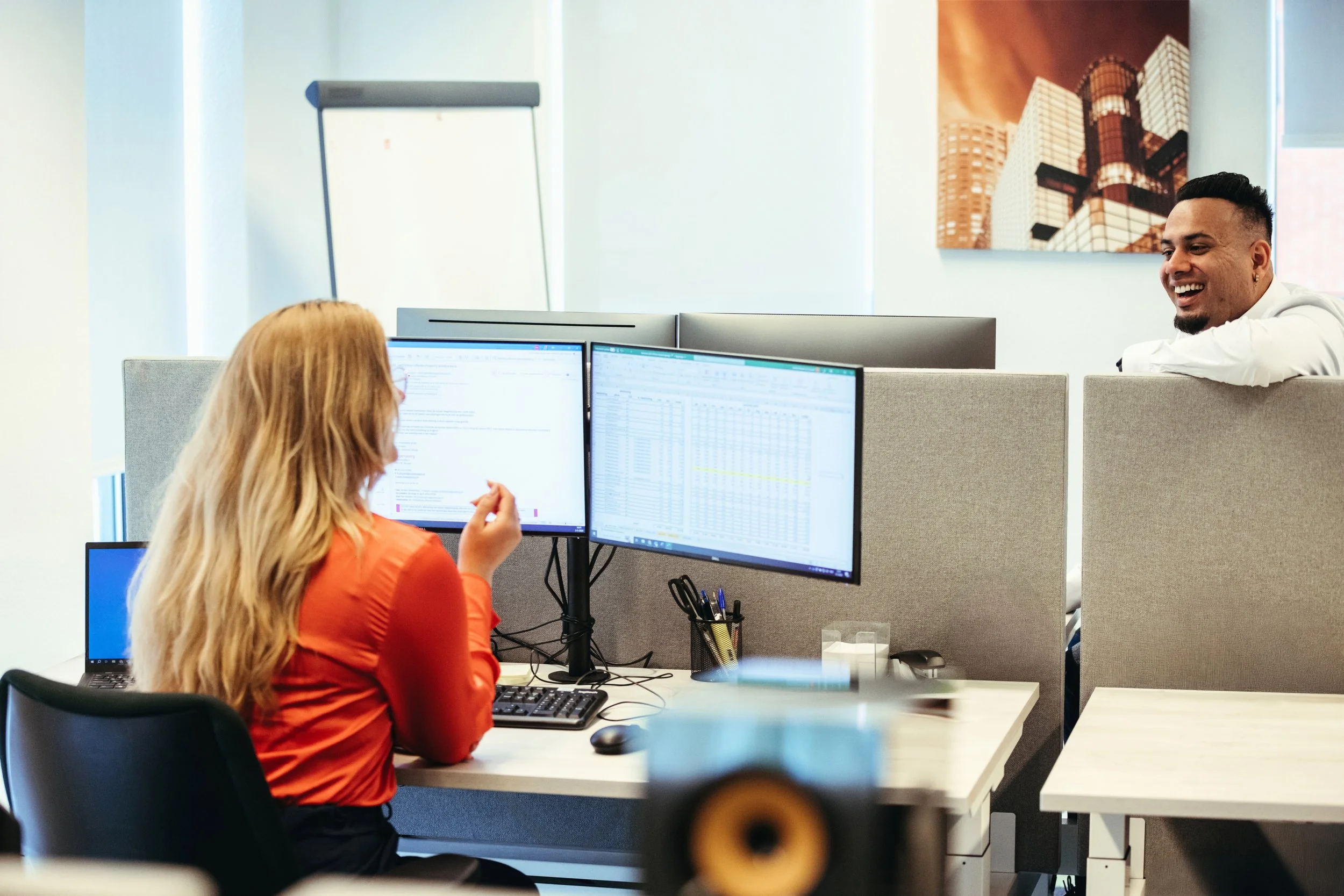 A woman with long blond hair and an orange shirt working at a dual-monitor computer setup in an office. She is looking at the screens and talking. A man with short black hair, white shirt, and beard is leaning on a partition, smiling at her. Office cubicles, a large window, and a cityscape picture on the wall are visible in the background.