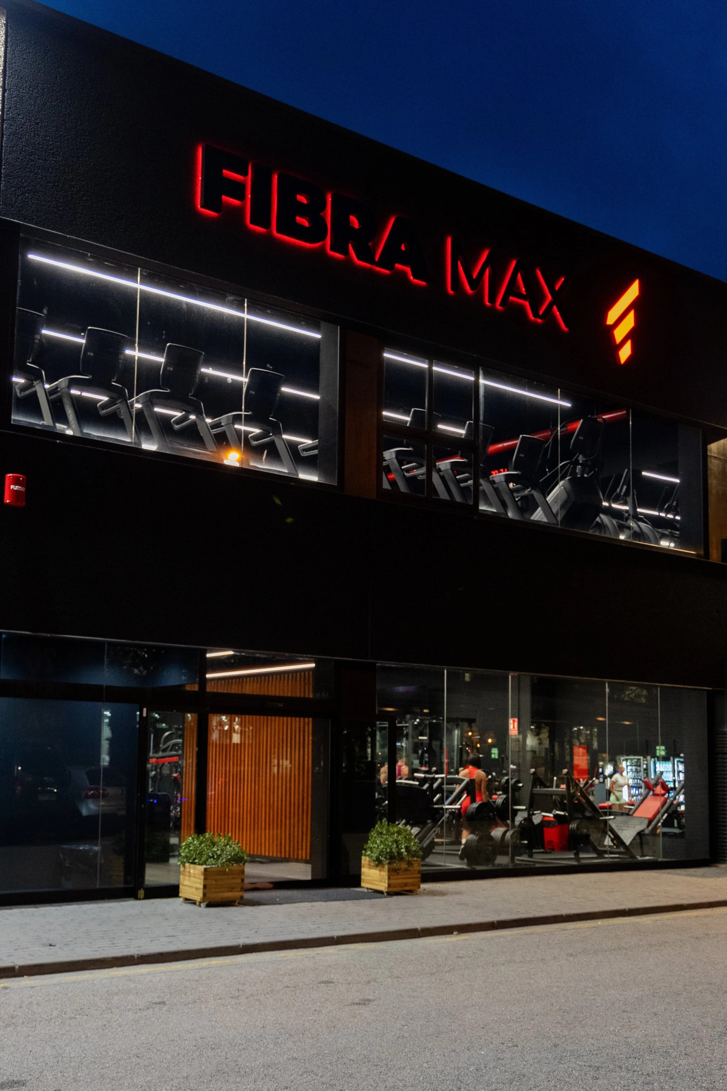 Vista frontal de una planta de gimnasio llamada 'FIBRA MAX' en la noche, con ventanas grandes que muestran máquinas de ejercicio y áreas de entrenamiento con personas entrenando.