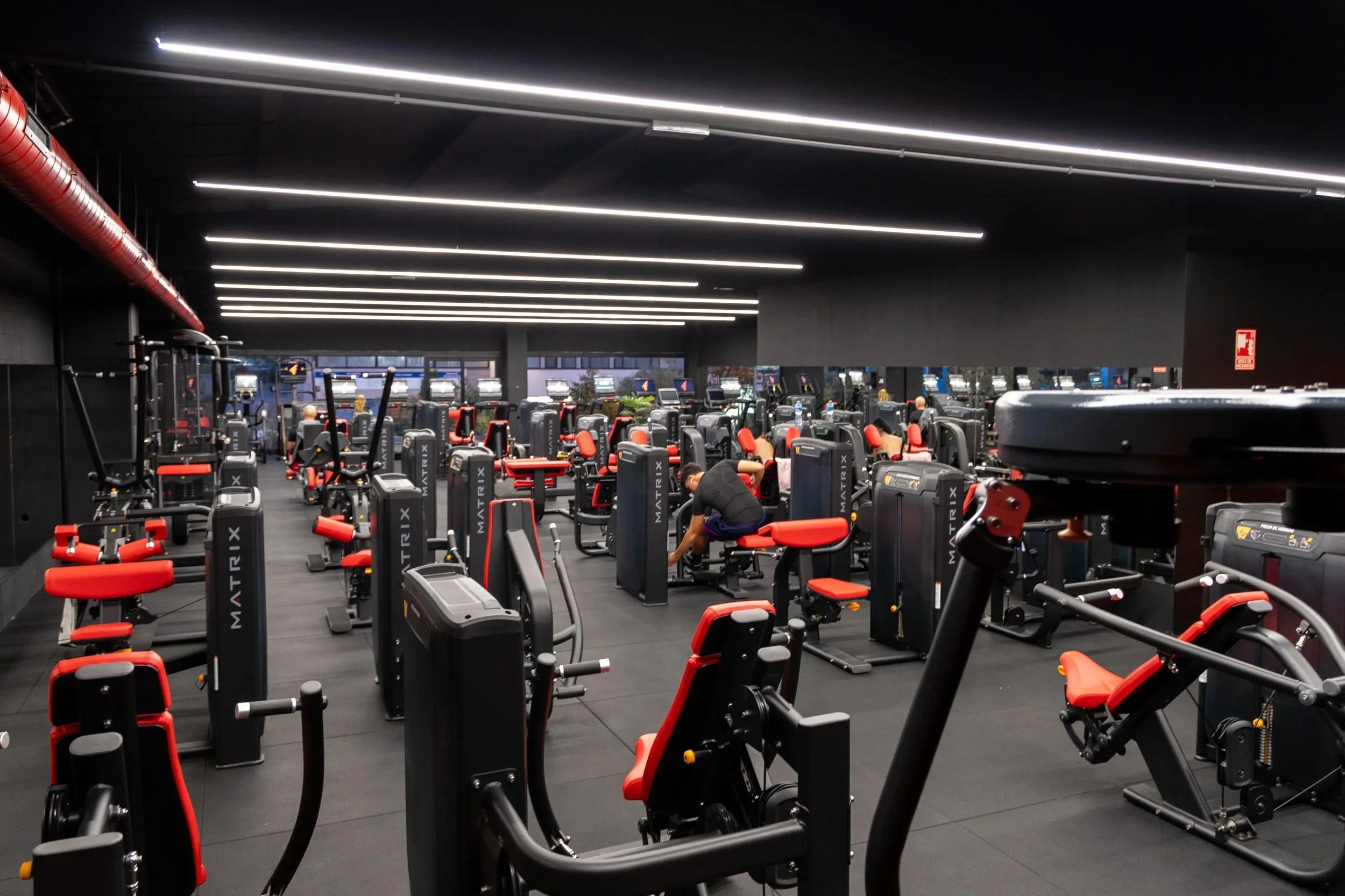 Piso de gimnasio con máquinas de ejercicio modernas de color negro y rojo, varias personas haciendo ejercicio, en un espacio amplio con techos negros y luces LED.