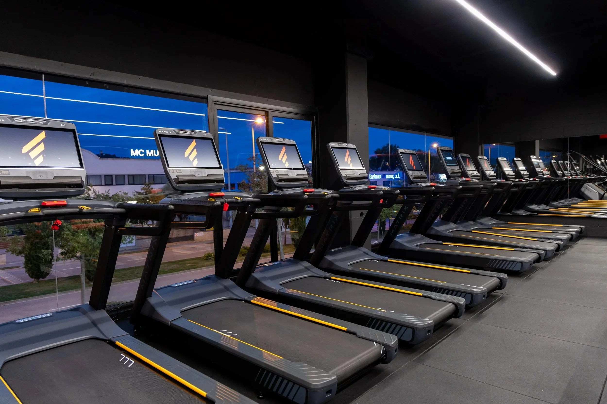 Cintas de correr en un gimnasio, en interiores, con pantallas digitales en la parte superior y ventanas que muestran la ciudad en la noche.