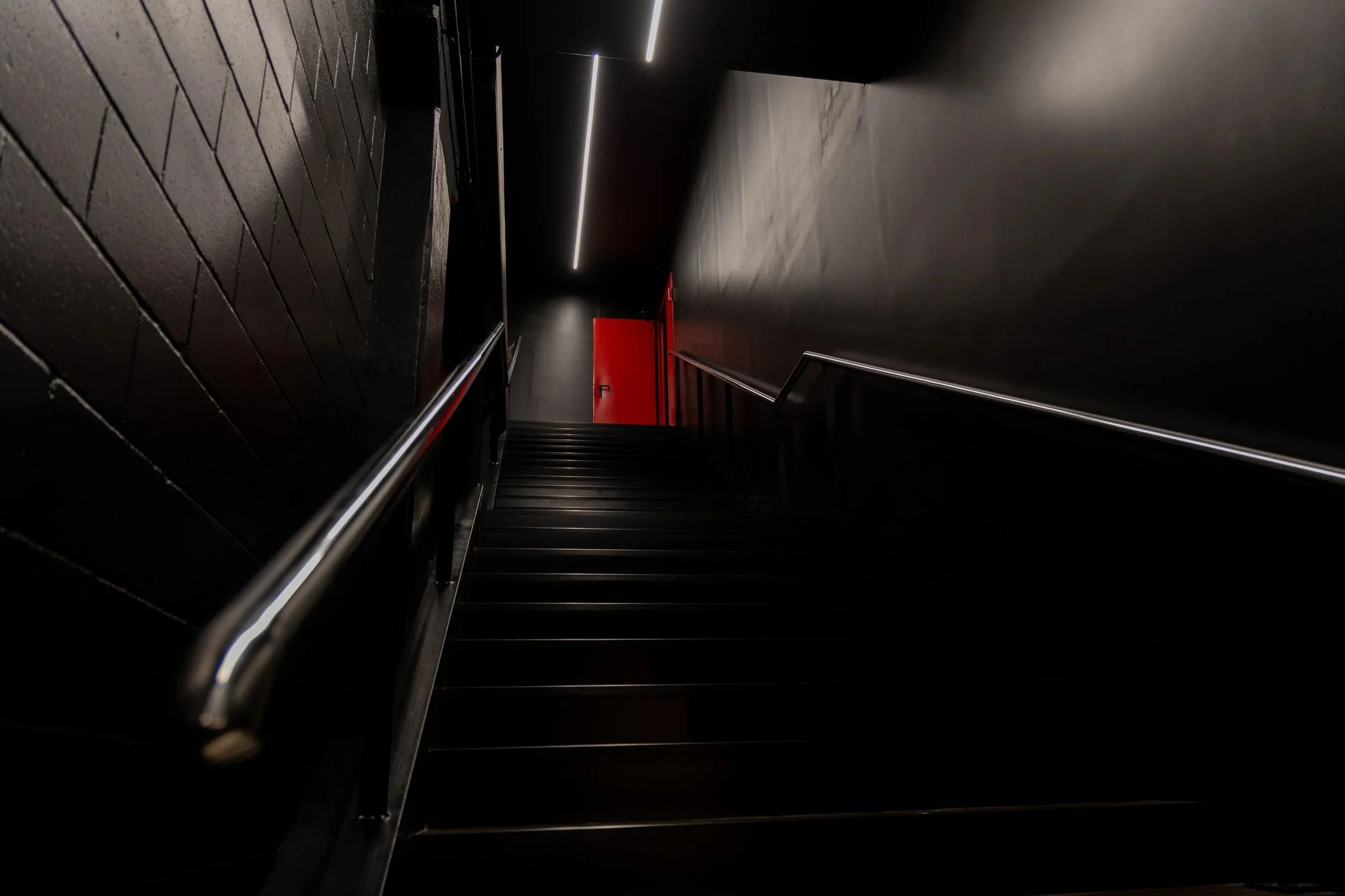 Escalera negra con pasamanos de metal, paredes oscuras y una puerta roja al fondo en un entorno moderno y minimalista.