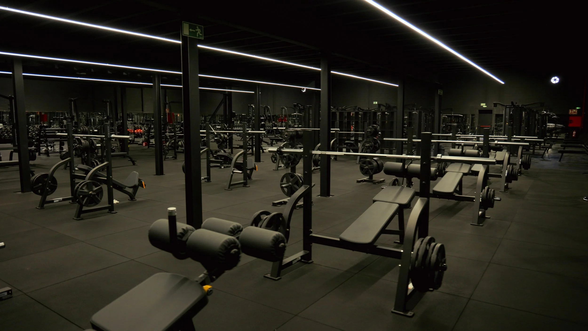 Interior de un gimnasio con equipos de ejercicio de color negro y pisos de goma negra, iluminado con luces blancas en el techo. Hay una señal de emergencia y algunas pesas apiladas en el fondo.