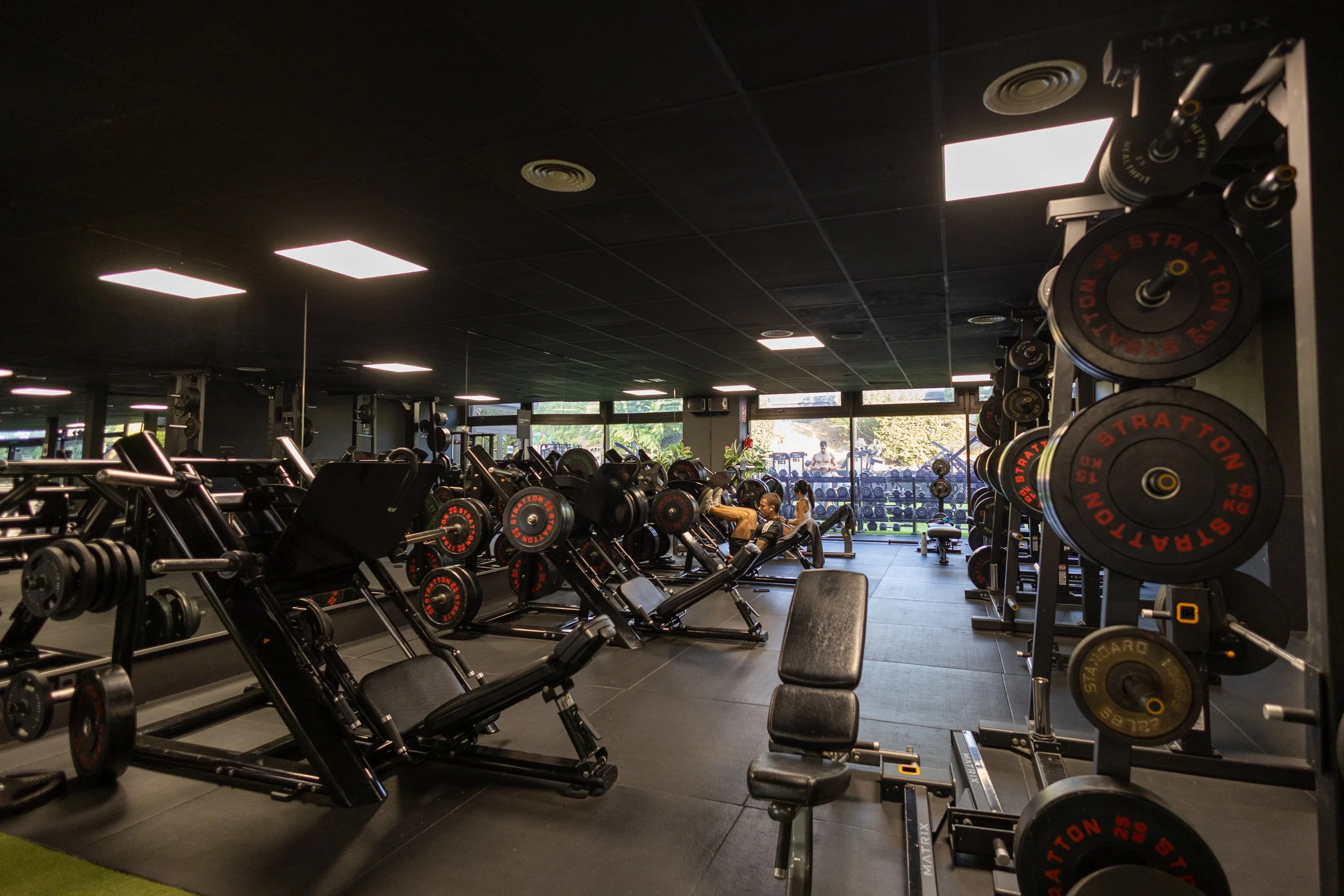 Interior de gimnasio con máquinas de pesas y área de entrenamiento, ventanas amplias y personas haciendo ejercicio.